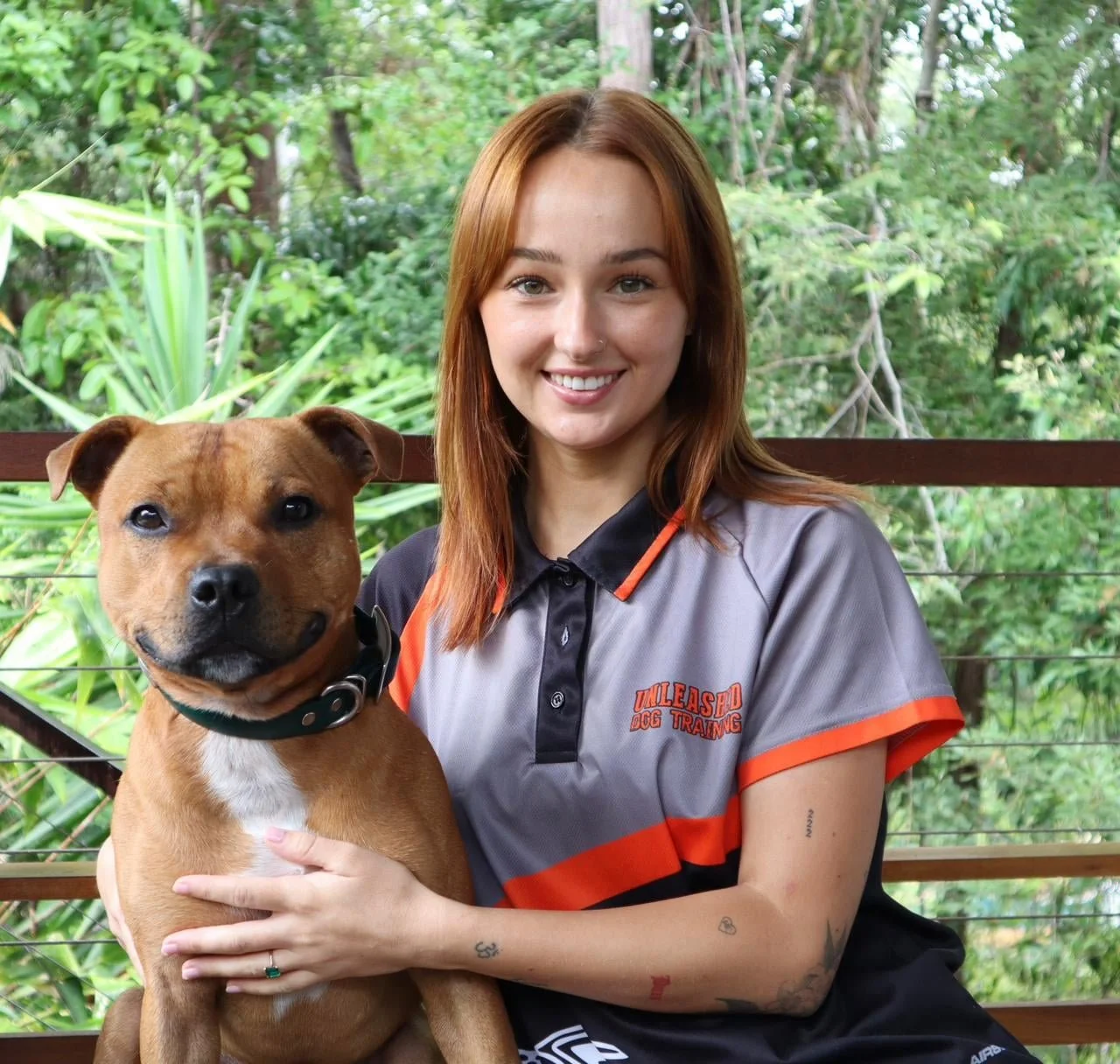 Woman with brown hair wearing a Unleashed Dog Training shirt, sitting outdoors with a brown dog.