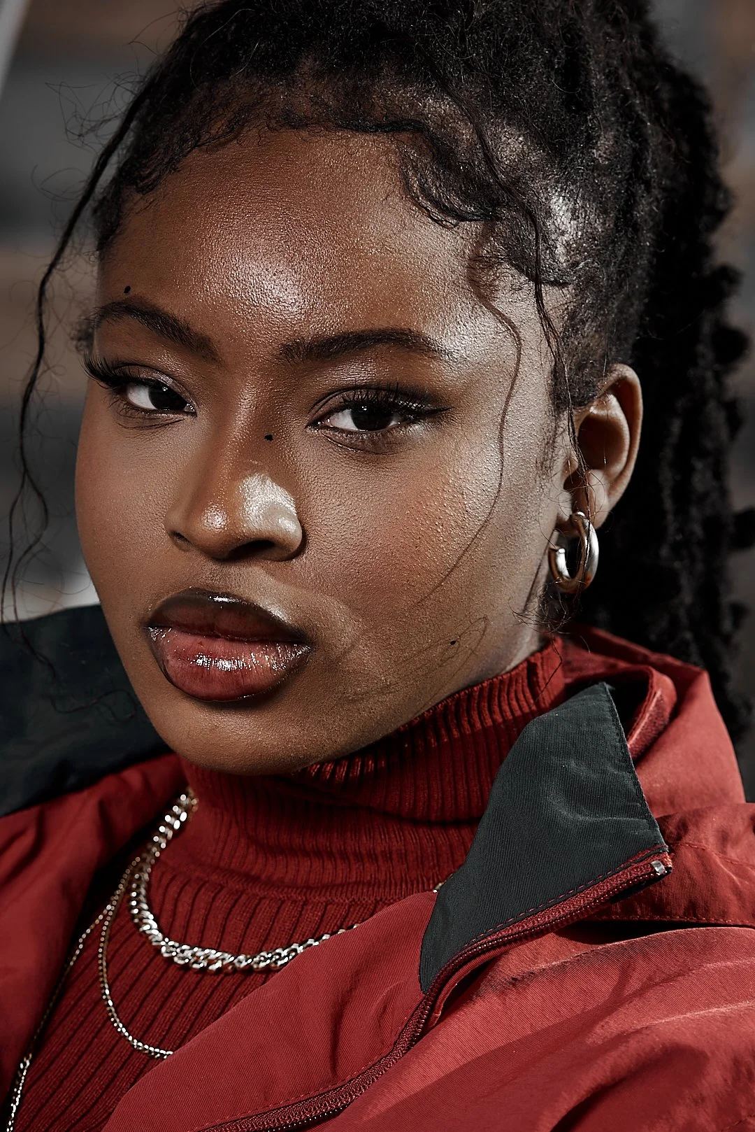 Close-up of a person with braided hair, wearing a red jacket and turtleneck, accessorized with hoop earrings and chain necklaces.