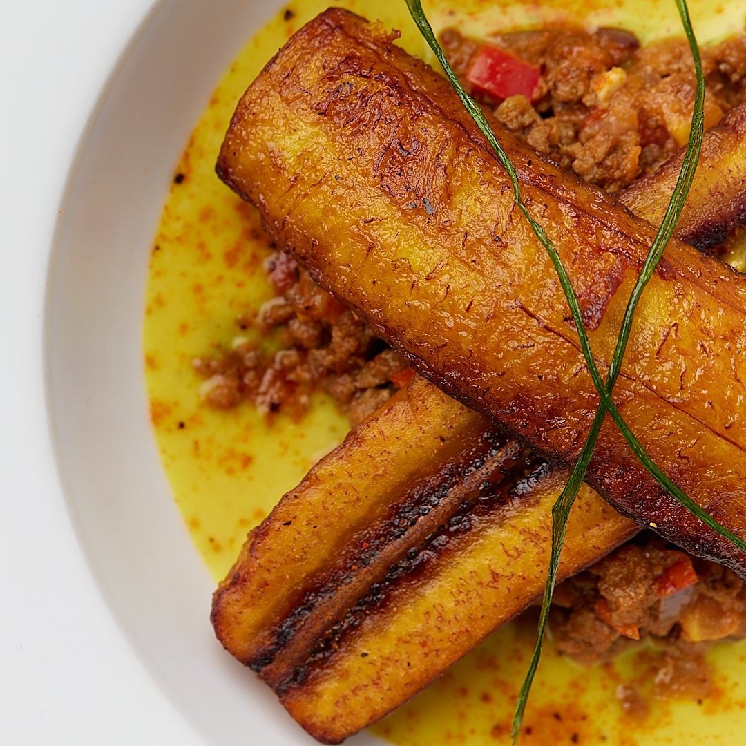 A dish featuring fried plantains with minced meat on a yellow sauce, garnished with a thin green herb.