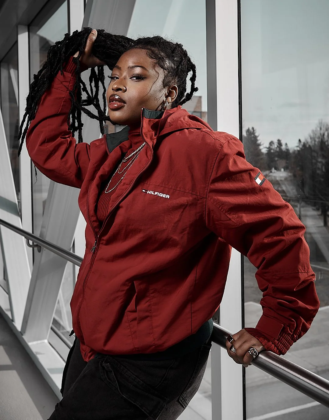Person wearing a red jacket with "Hilfiger" logo, standing indoors, holding their braided hair, and looking at the camera.