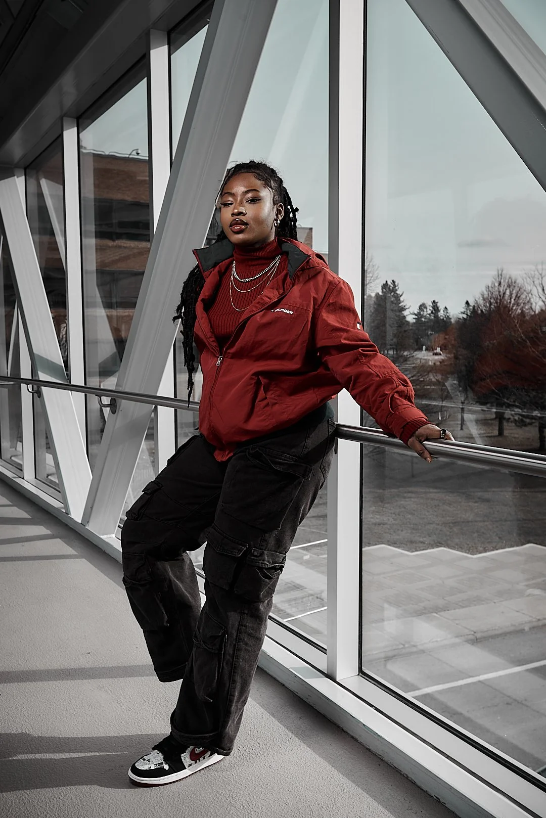 A person wearing a red jacket and black cargo pants leans against a railing in a glass-walled corridor, with an outdoor scene visible through the windows.