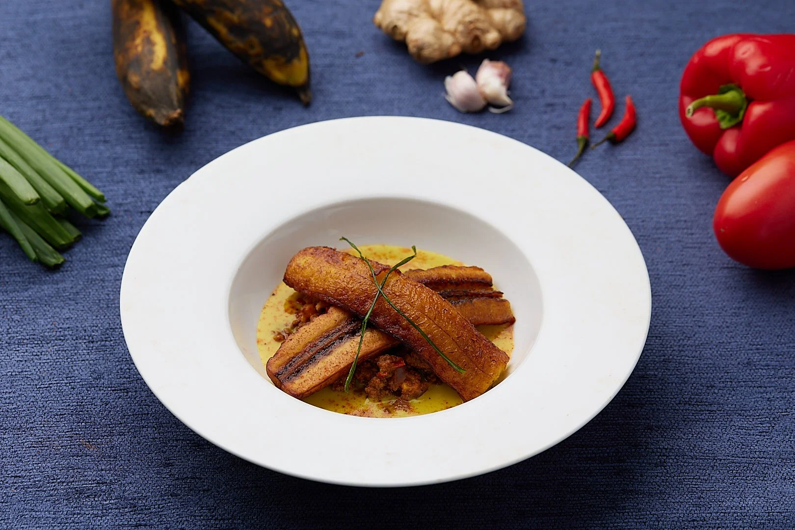 A plate of fried plantains with sauce on a blue tablecloth, surrounded by fresh ingredients such as green onions, ripe plantains, ginger, garlic, hot peppers, and red bell peppers.