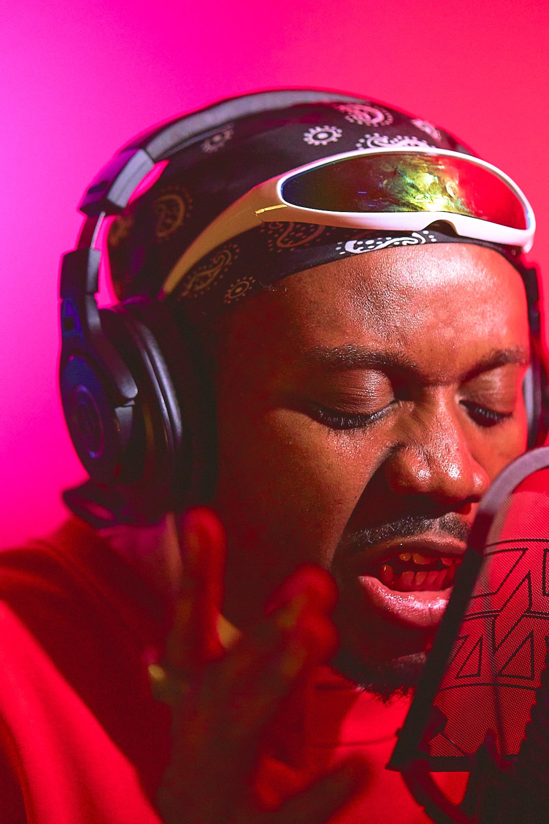 A person with glasses and a headscarf wearing headphones, singing into a microphone with a red background.