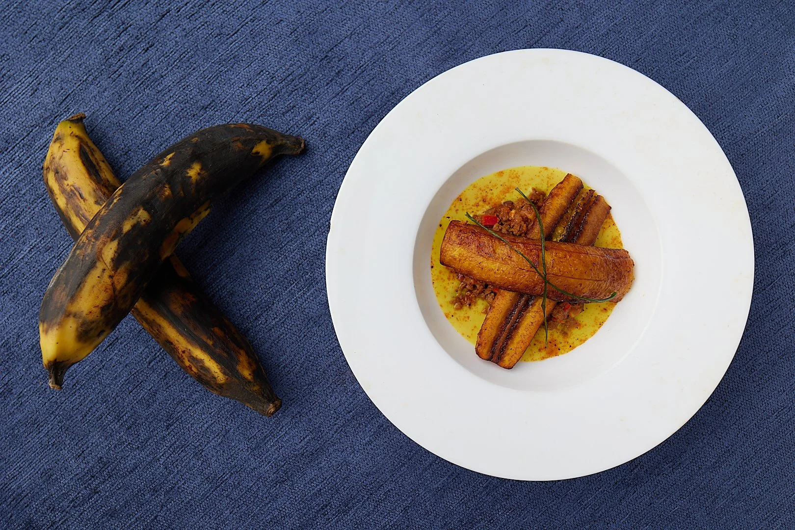 Fried plantains on a white plate with yellow sauce, next to two ripe plantains on a blue textured surface.