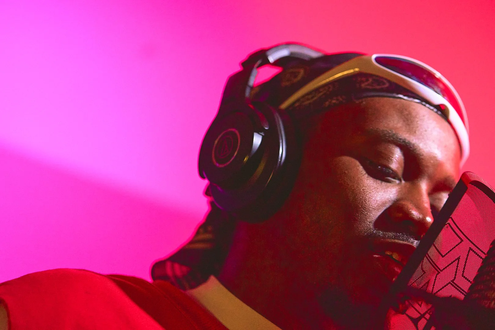 Person wearing headphones and a bandana, speaking into a microphone with a pop filter, against a pink background.