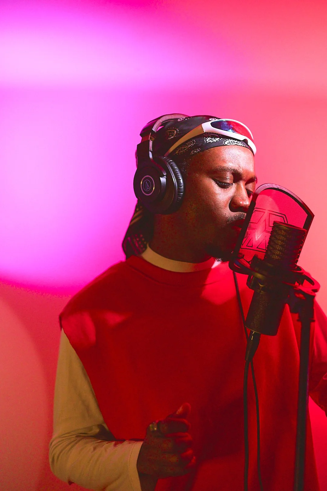 Person singing into a microphone in a recording studio, wearing headphones and a headscarf, with a pink and red background.