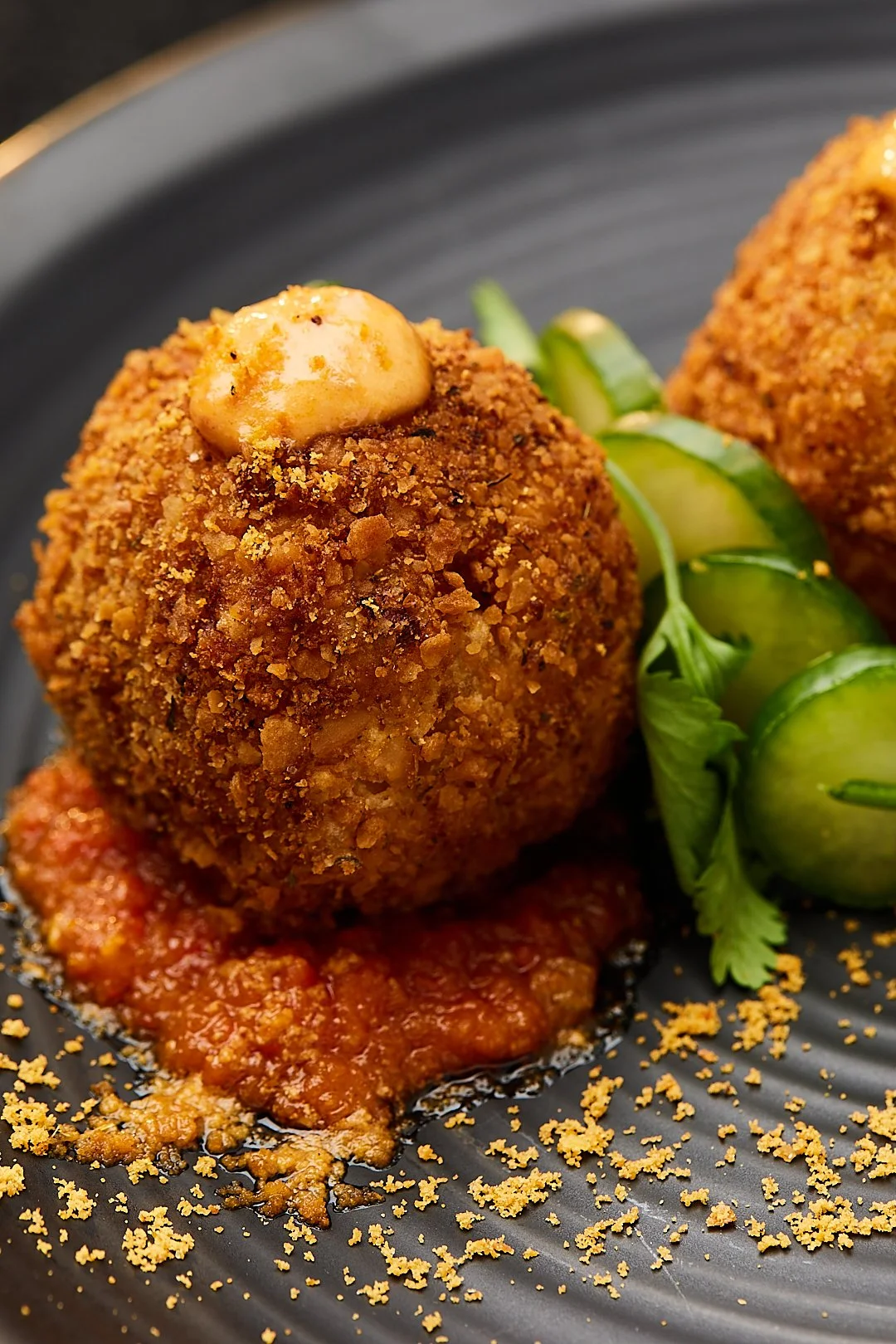Close-up of a crispy arancini ball with creamy sauce on top, served on a black plate with tomato sauce, garnished with sliced cucumbers and herbs.
