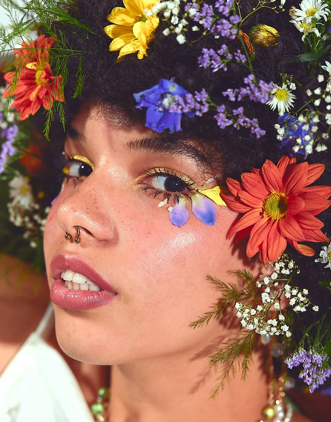 Close-up of a person with curly hair adorned with colorful flowers, including yellow, purple, red, and white blooms. They have petal decorations under their eye and wear a nose ring.