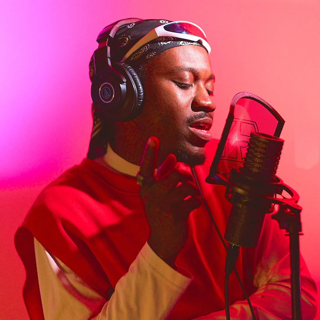 Man wearing headphones and a red shirt, singing into a microphone in a recording studio, with colored lighting in the background.