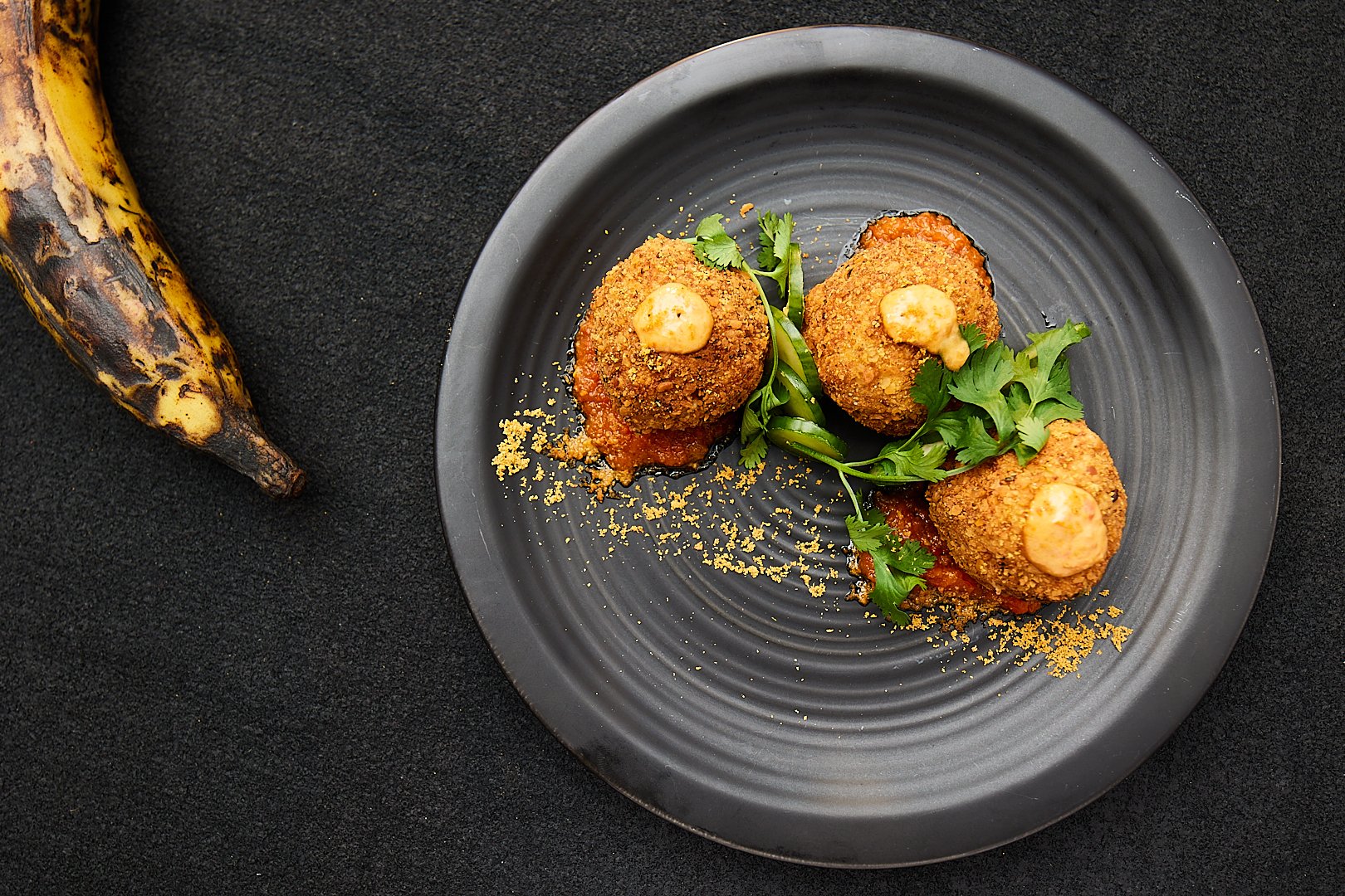 Three deep-fried balls on a black plate garnished with sauce and herbs, next to a ripe plantain on a black background.
