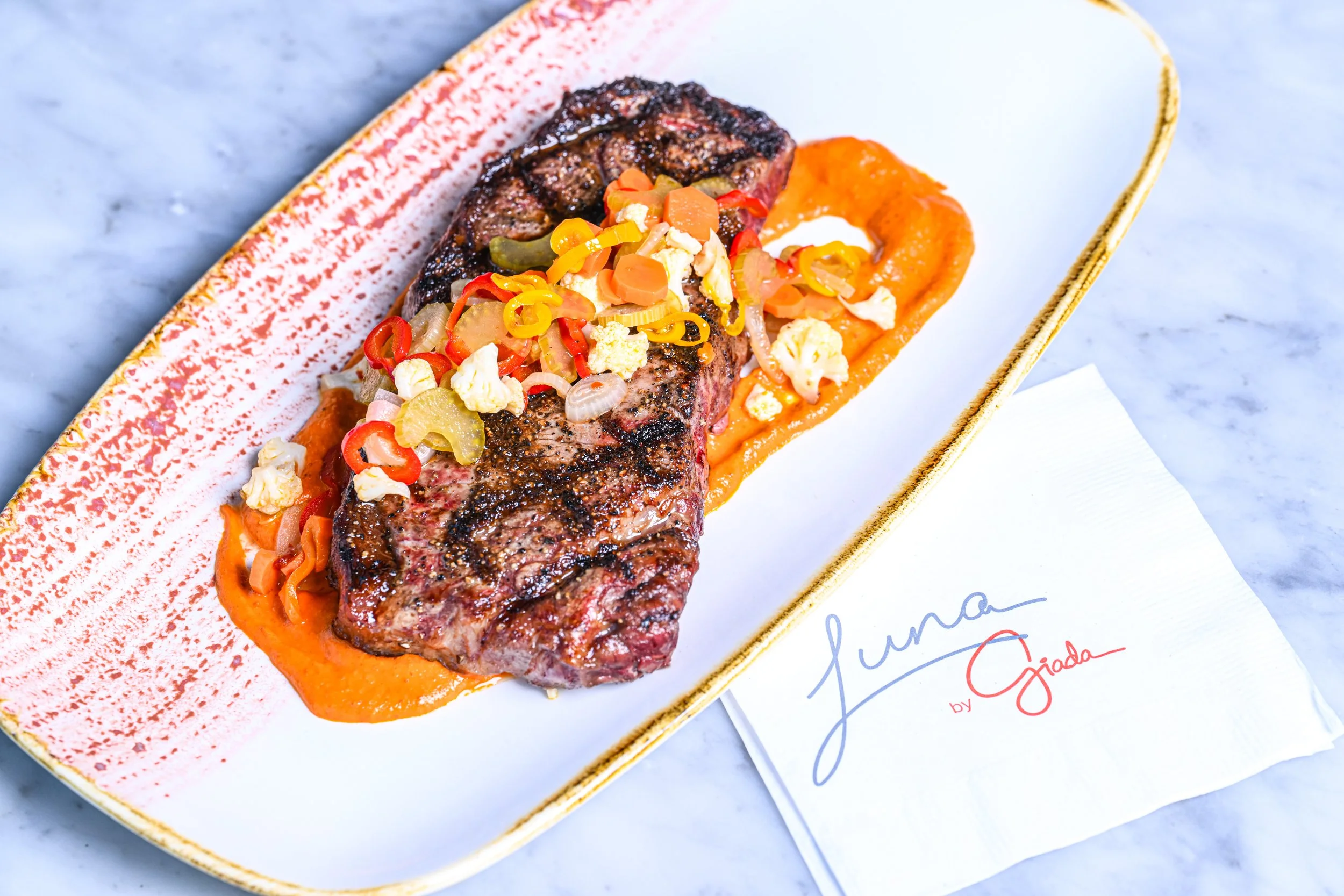 A grilled steak topped with diced vegetables and crumbled cheese on a bed of orange sauce, served on a decorative rectangular plate with a white paper underneath.