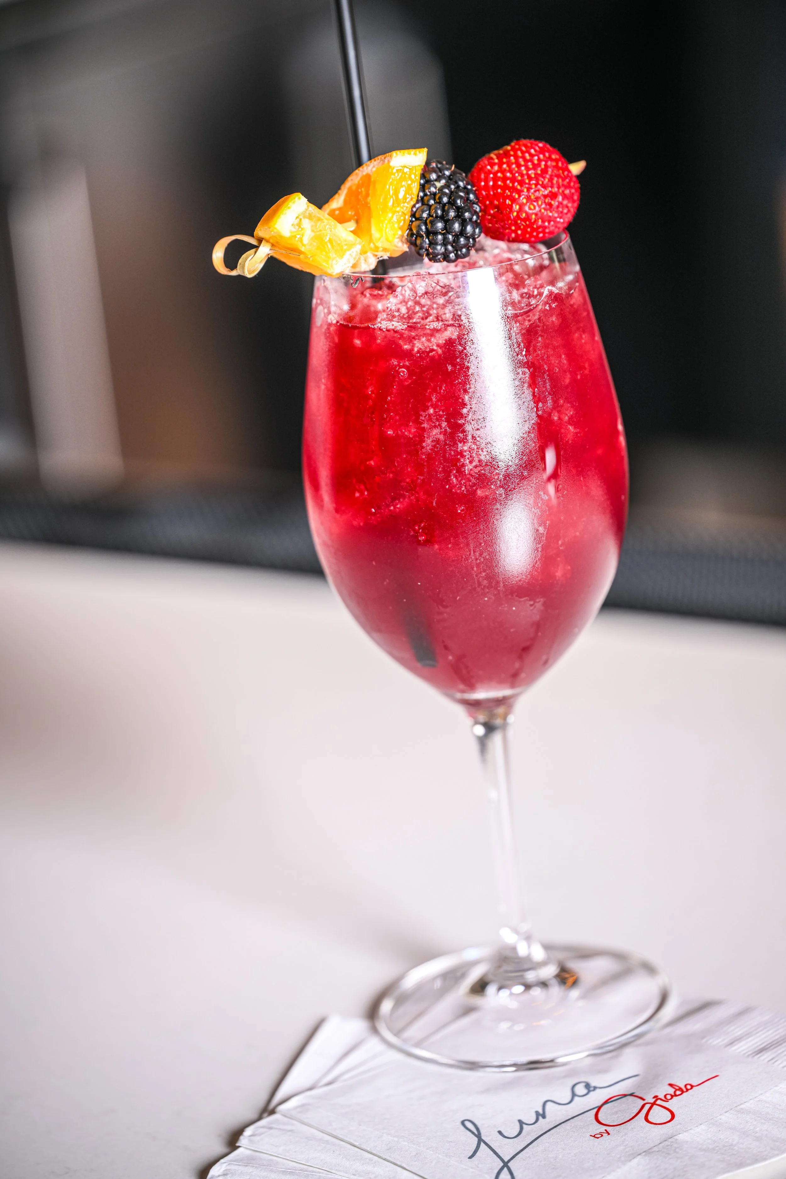 A red cocktail garnished with orange slices, blackberries, and strawberries in a wine glass, placed on a white napkin.