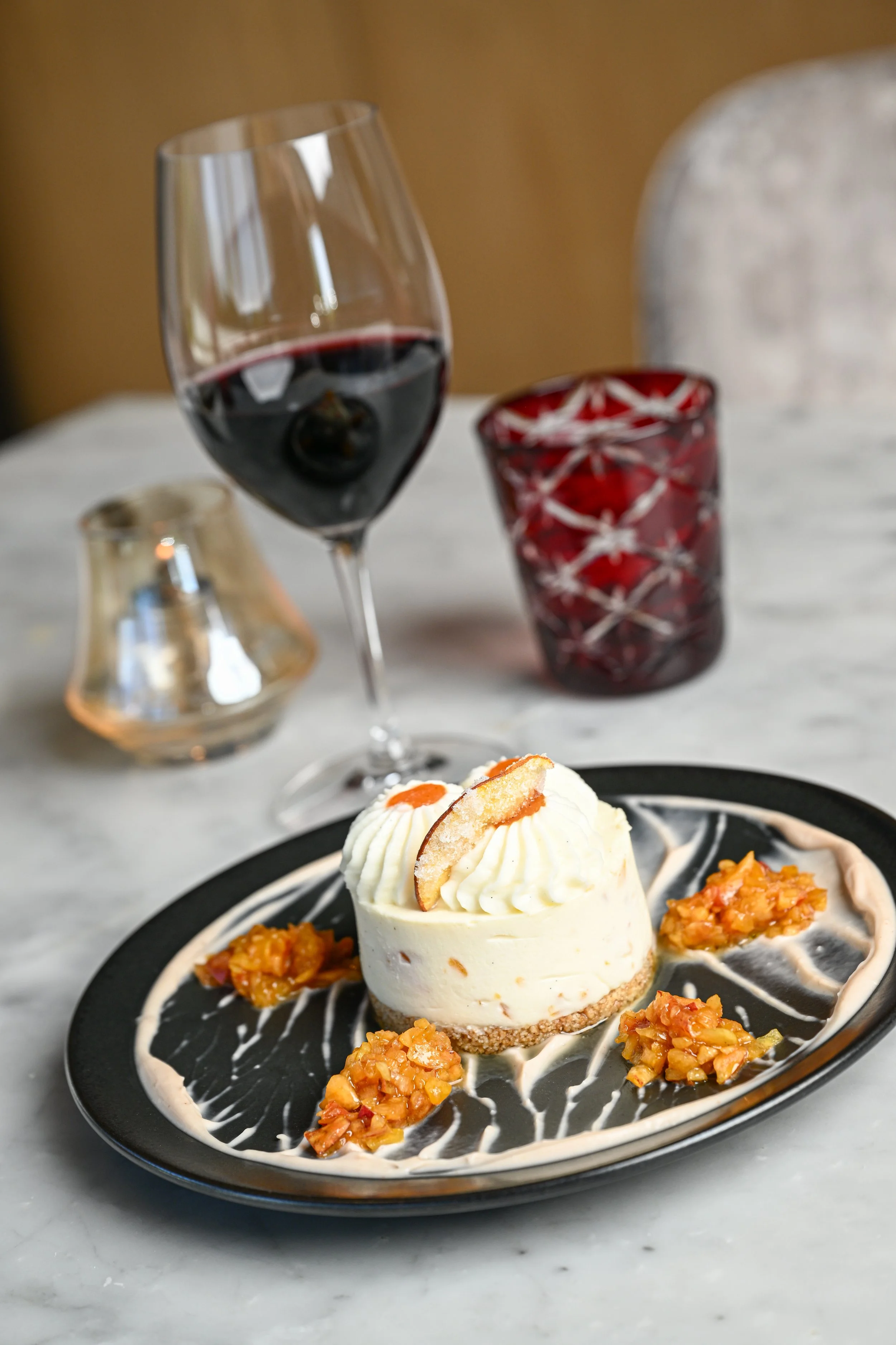 A dessert with a creamy cake topped with a cookie slice on a black plate, accompanied by four spoonfuls of fruit compote, and a glass of red wine on a white marble table.