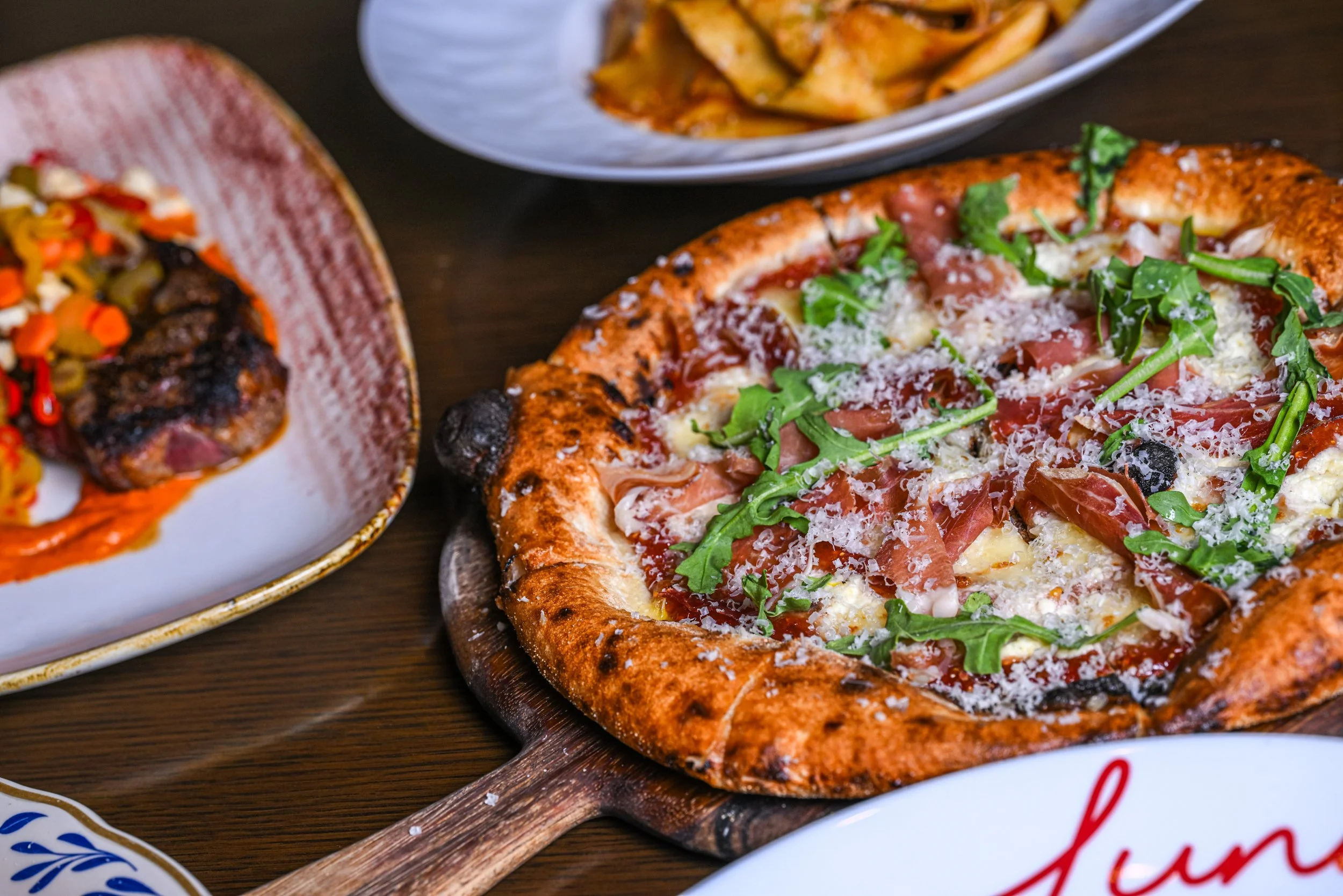 A pizza with arugula, ham, cheese, and tomato slices on a wooden board, with other plates of pasta and meat in the background.