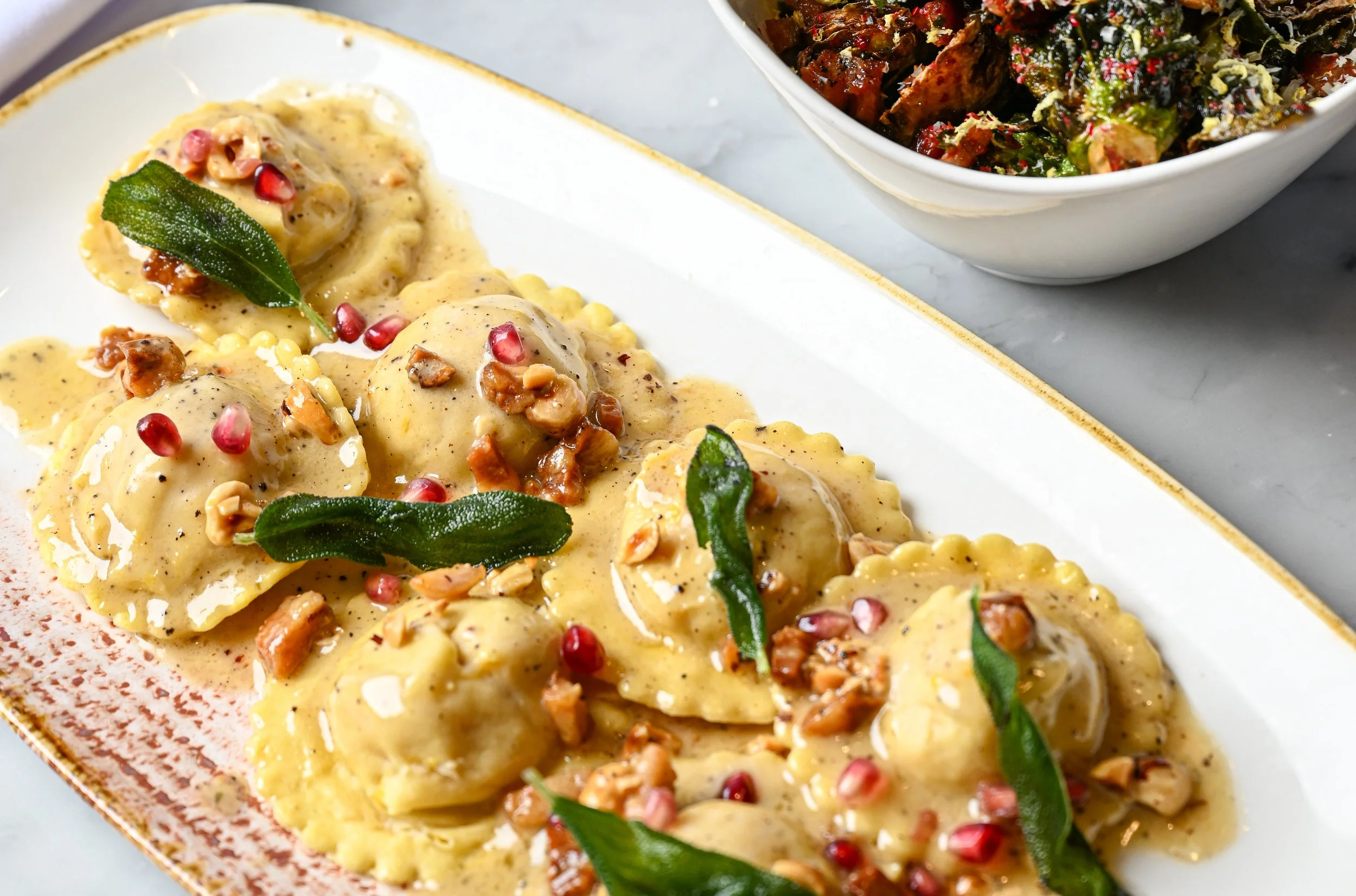 Plate of ravioli topped with creamy sauce, garnished with green herbs, pomegranate seeds, and chopped nuts. In the background, a bowl of chopped vegetable salad is visible.