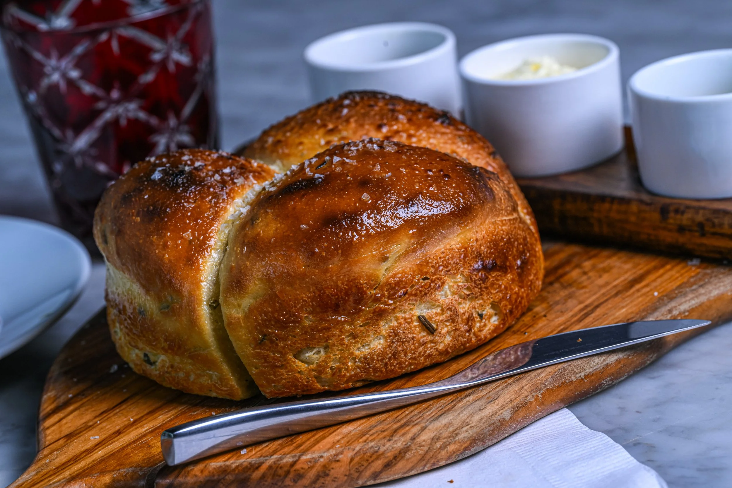 A freshly baked loaf of bread with a golden-brown crust on a wooden cutting board, surrounded by small white bowls of dipping sauces, with a glass of dark-colored beverage in the background.