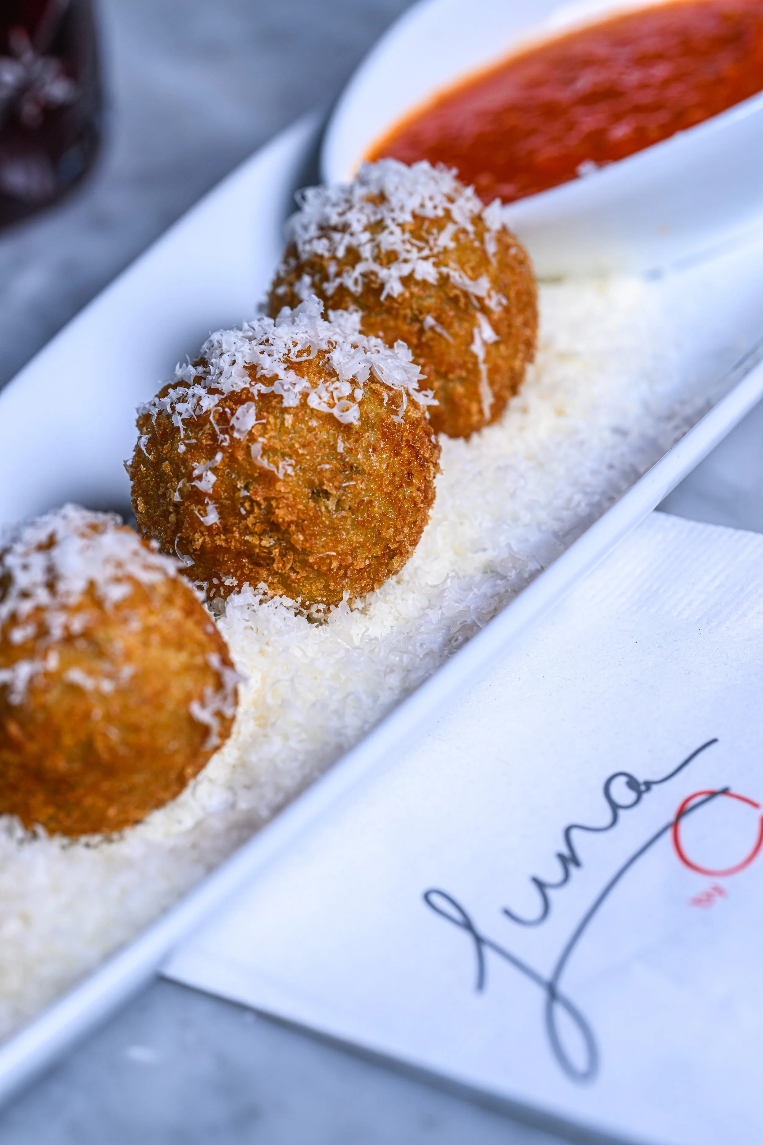 Close-up of three breaded and fried croquettes topped with shredded cheese, served on a white plate with a small piece of breaded meat or fish in the background.