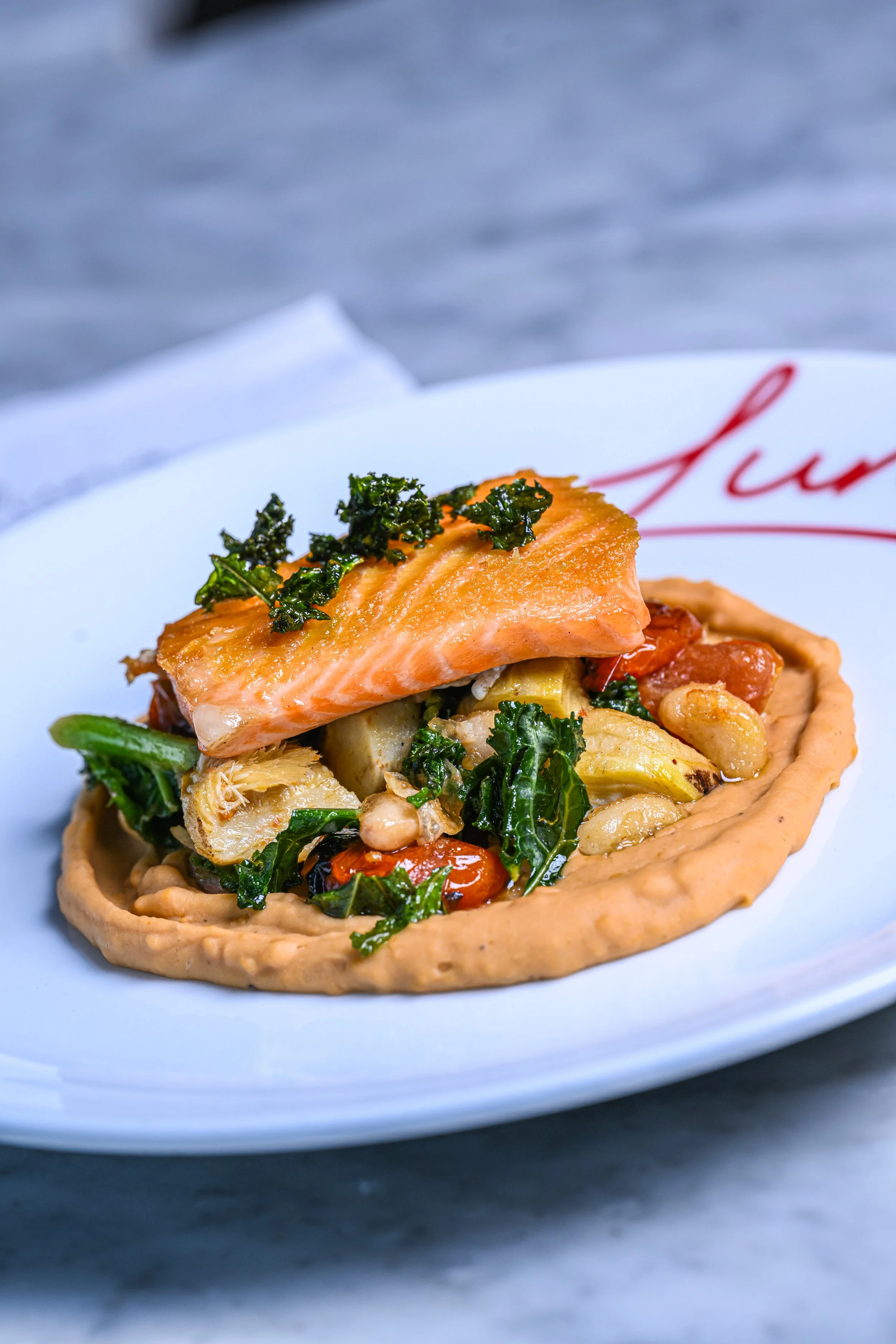 A savory tart filled with vegetables, including kale, cherry tomatoes, cauliflower, and topped with a cooked salmon fillet and herbs, served on a white plate with red writing in the background.