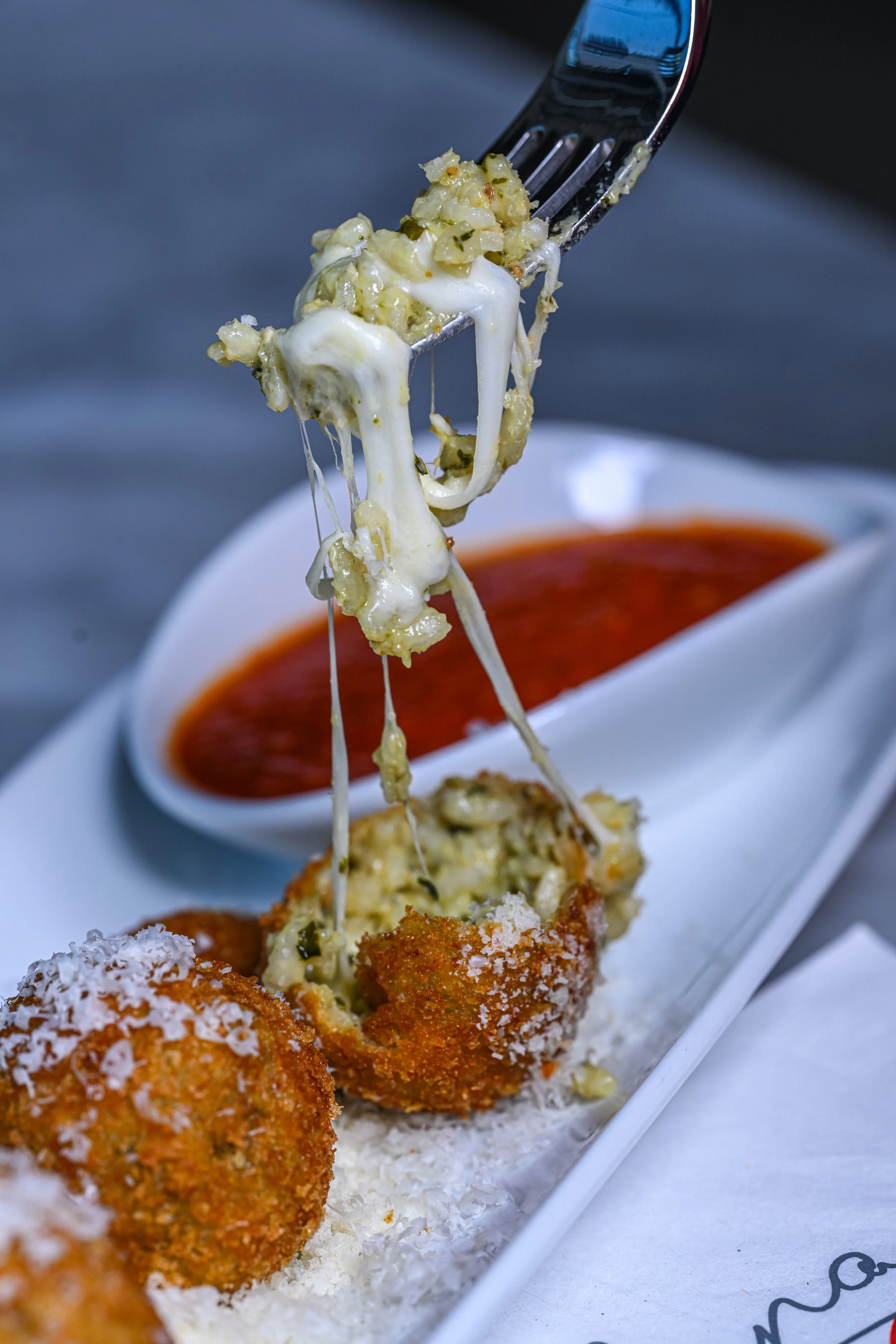 Close-up of a fork lifting cheesy, herb-seasoned stuffing from a breaded, fried ball of food, with a side of marinara sauce in a white dish in the background.