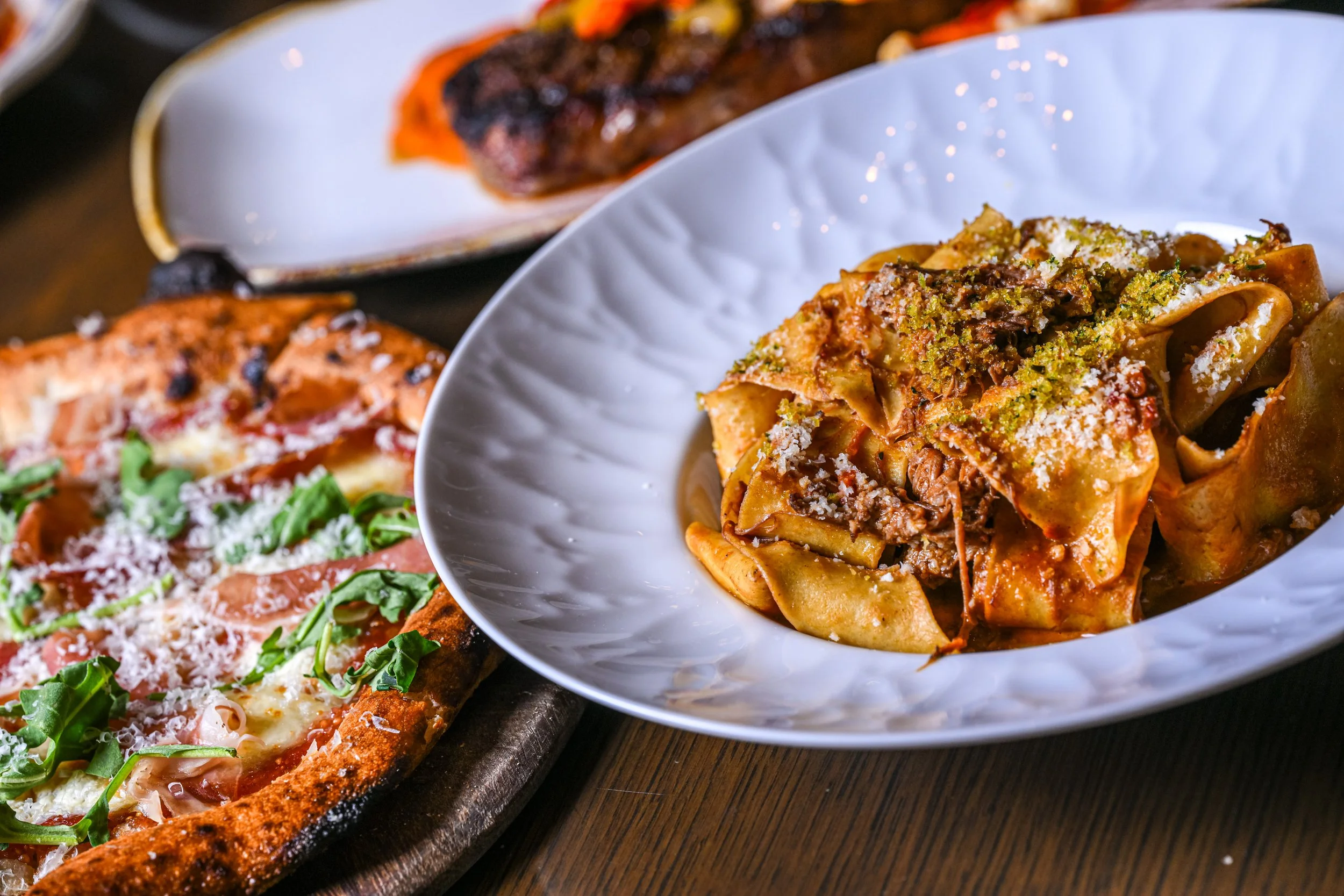 Close-up of pasta with meat sauce in a white bowl, with other dishes including pizza and grilled meat visible in the background.