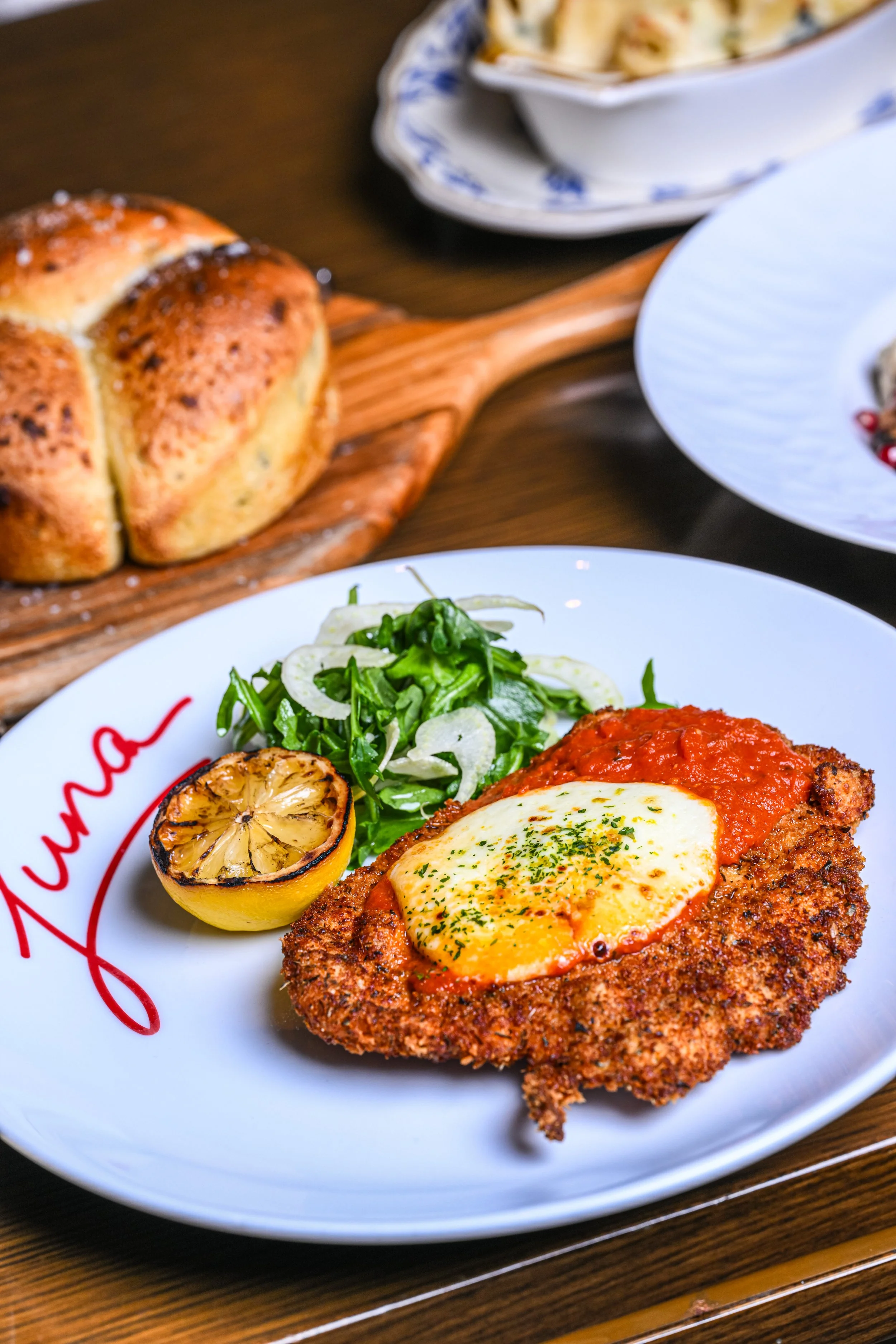 A plate of Italian food with a breaded veal cutlet topped with tomato sauce and melted cheese, garnished with parsley. Alongside are grilled lemon halves and a green salad. In the background, a loaf of bread and dishes of pasta are visible on a woode