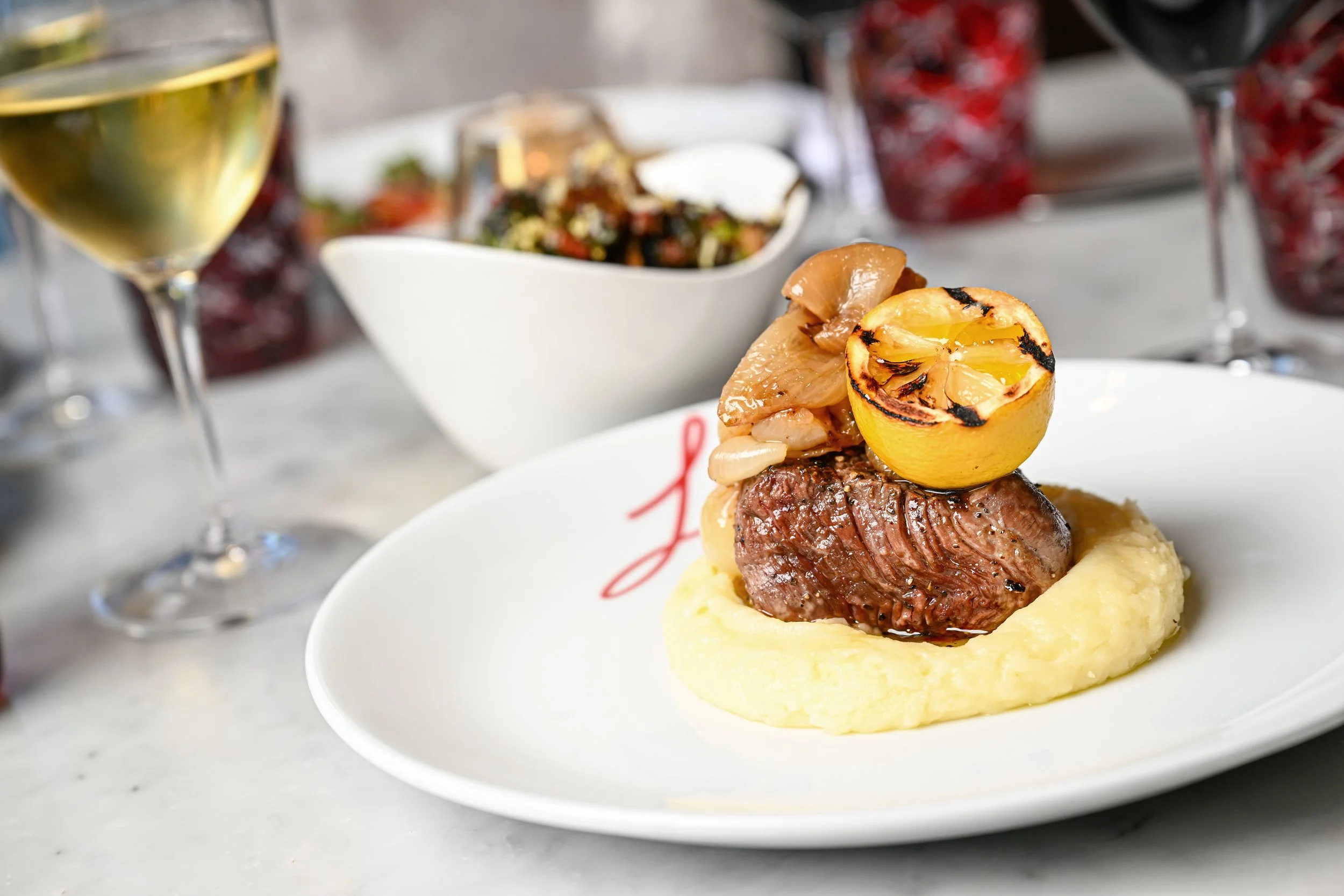 Filet mignon steak topped with grilled lemon, mushrooms, and onions, served on mashed potatoes, with glasses of white and red wine in the background.