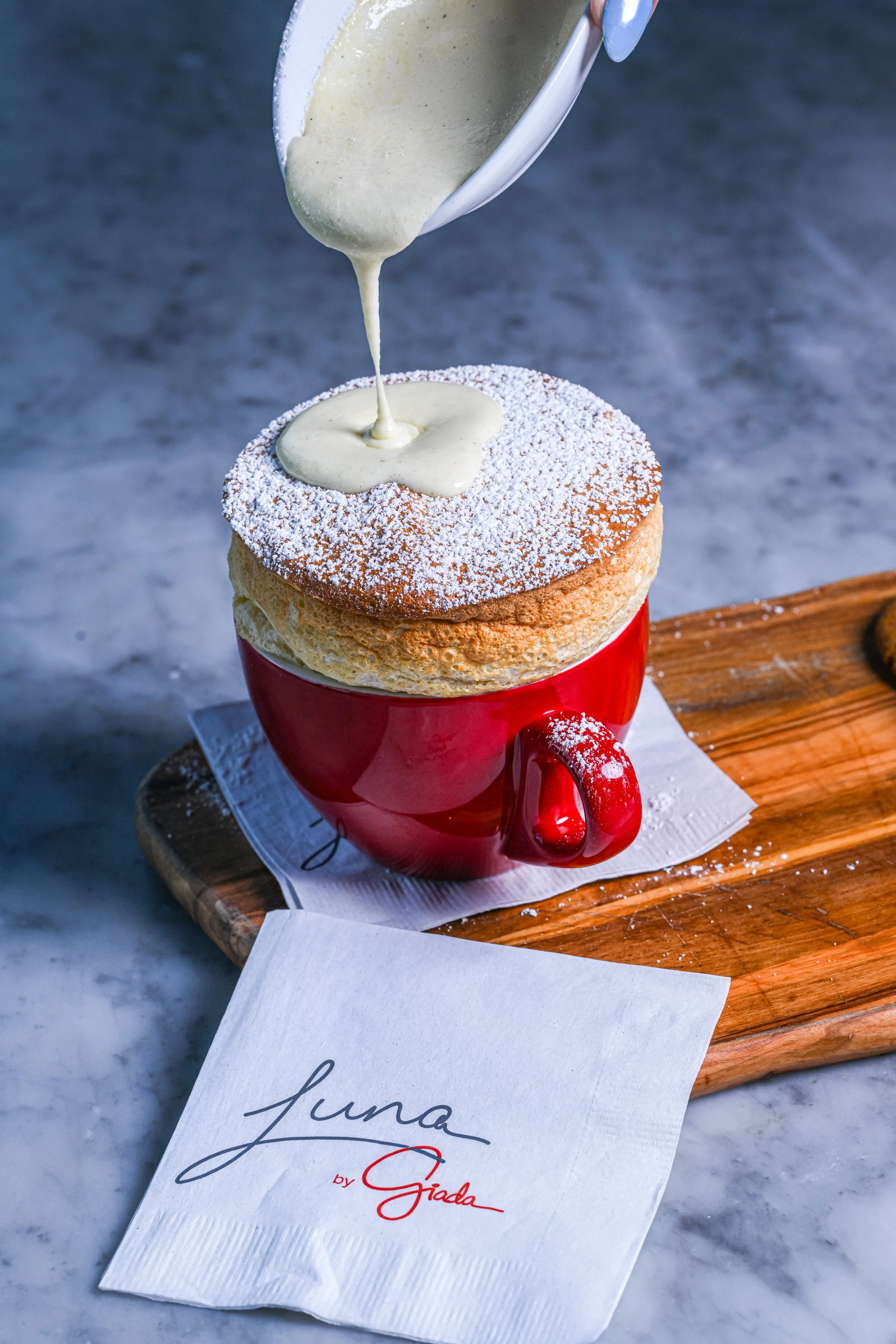 A cup of coffee topped with whipped cream and sprinkled with powdered sugar, placed on a wooden tray with a paper napkin underneath, and a piece of white paper with cursive writing partially visible in the foreground.
