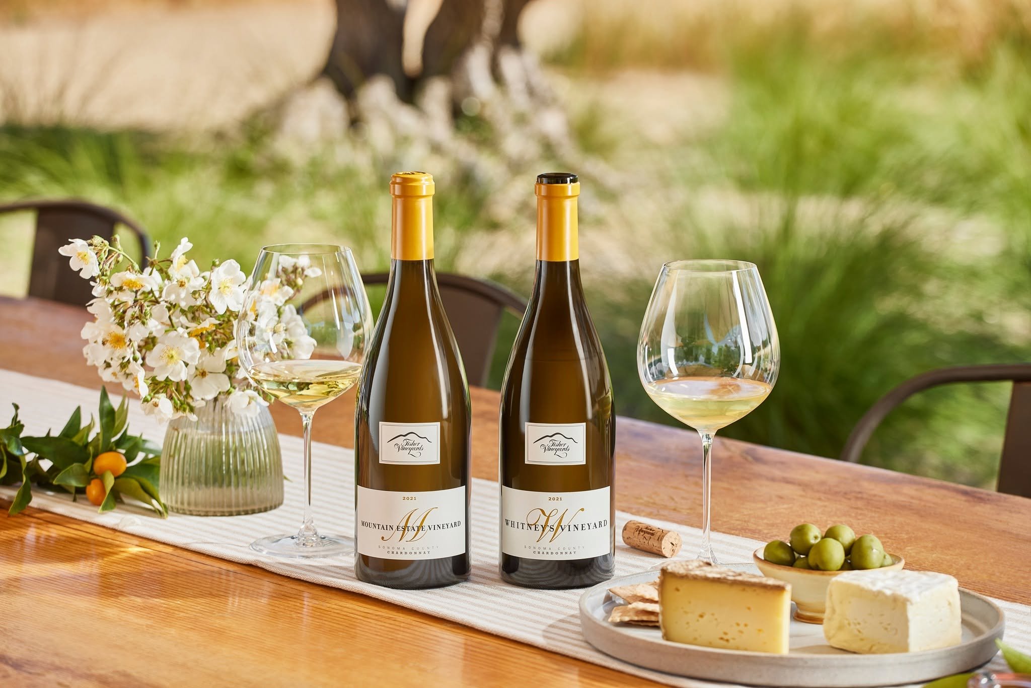 Two bottles of white wine, two wine glasses filled with white wine, a plate of cheese, grapes, and crackers, and a floral arrangement on a wooden table outdoors with greenery in the background.