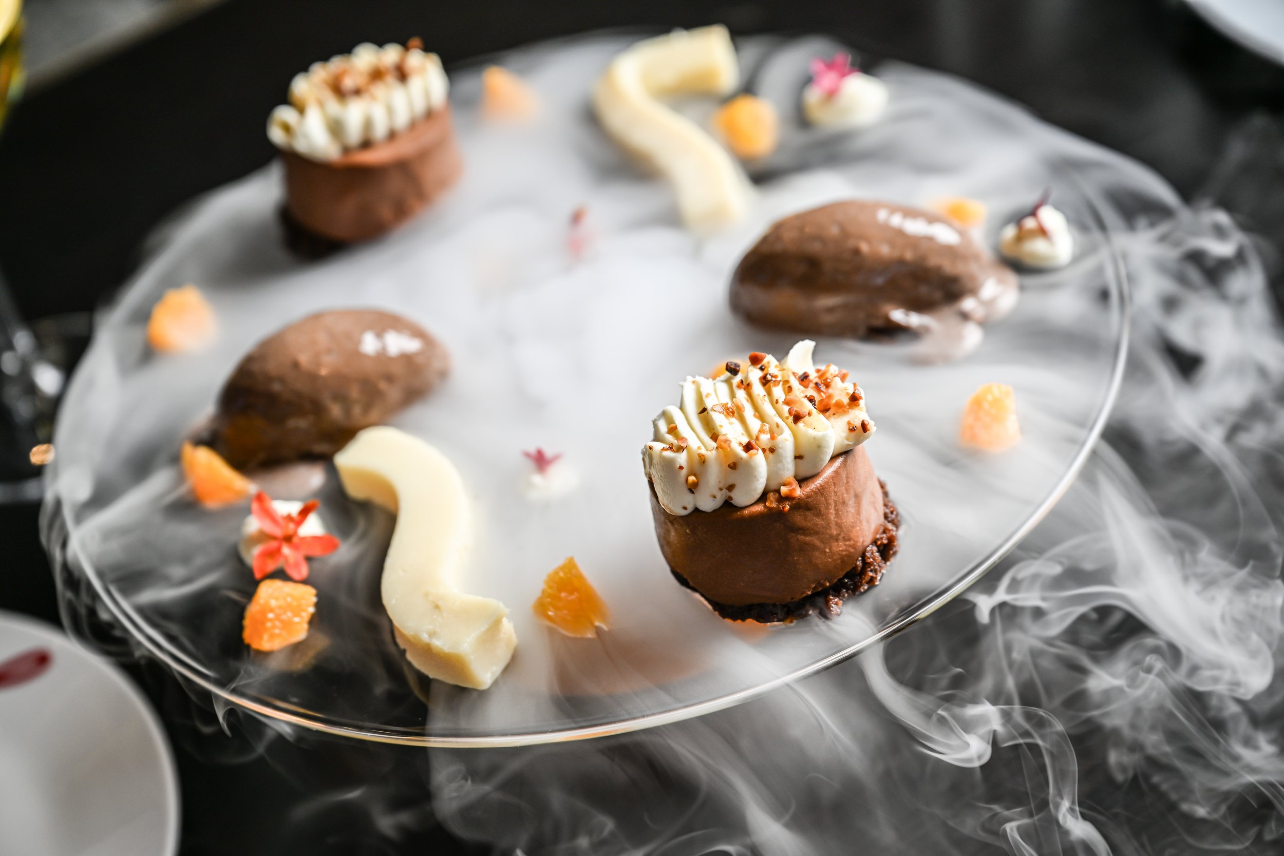Chocolate dessert with whipped cream topping on a glass plate, garnished with small edible flowers and fruit pieces, with smoke swirling around the plate.
