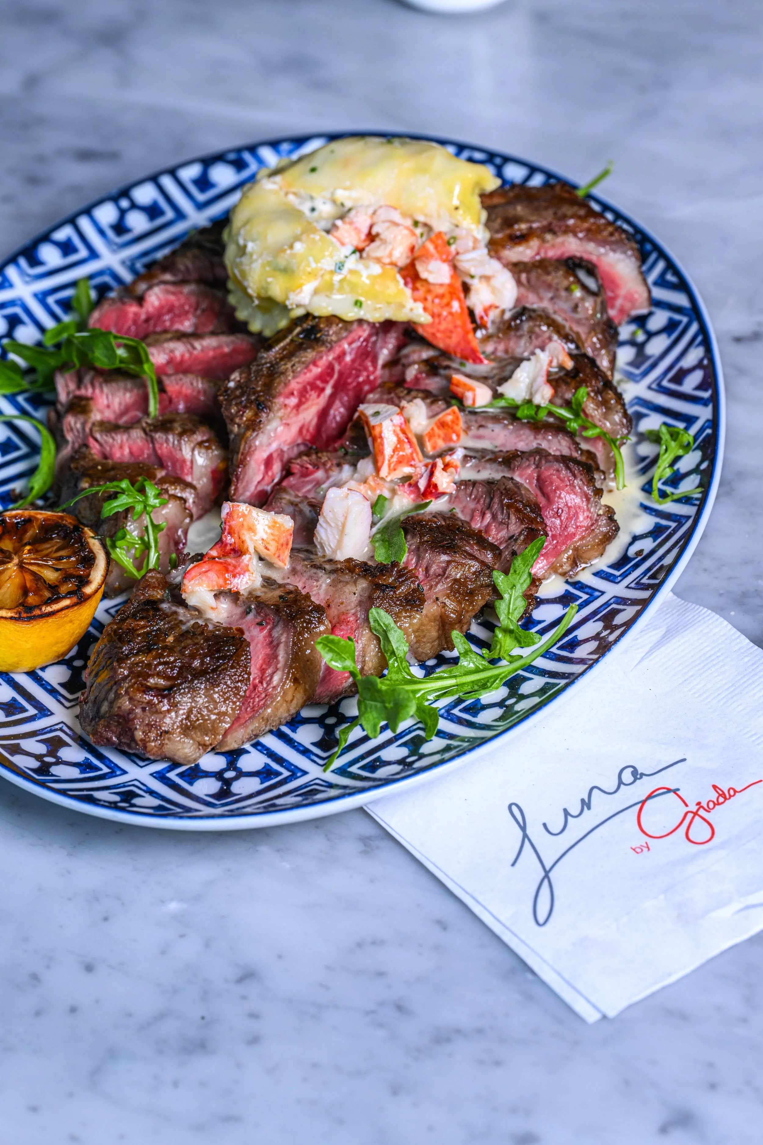 Sliced medium-rare steak topped with a dollop of creamy herb sauce, garnished with greens, served on a patterned blue and white plate, with a charred lemon half on a white marble surface, and a napkin with handwritten text in the background.