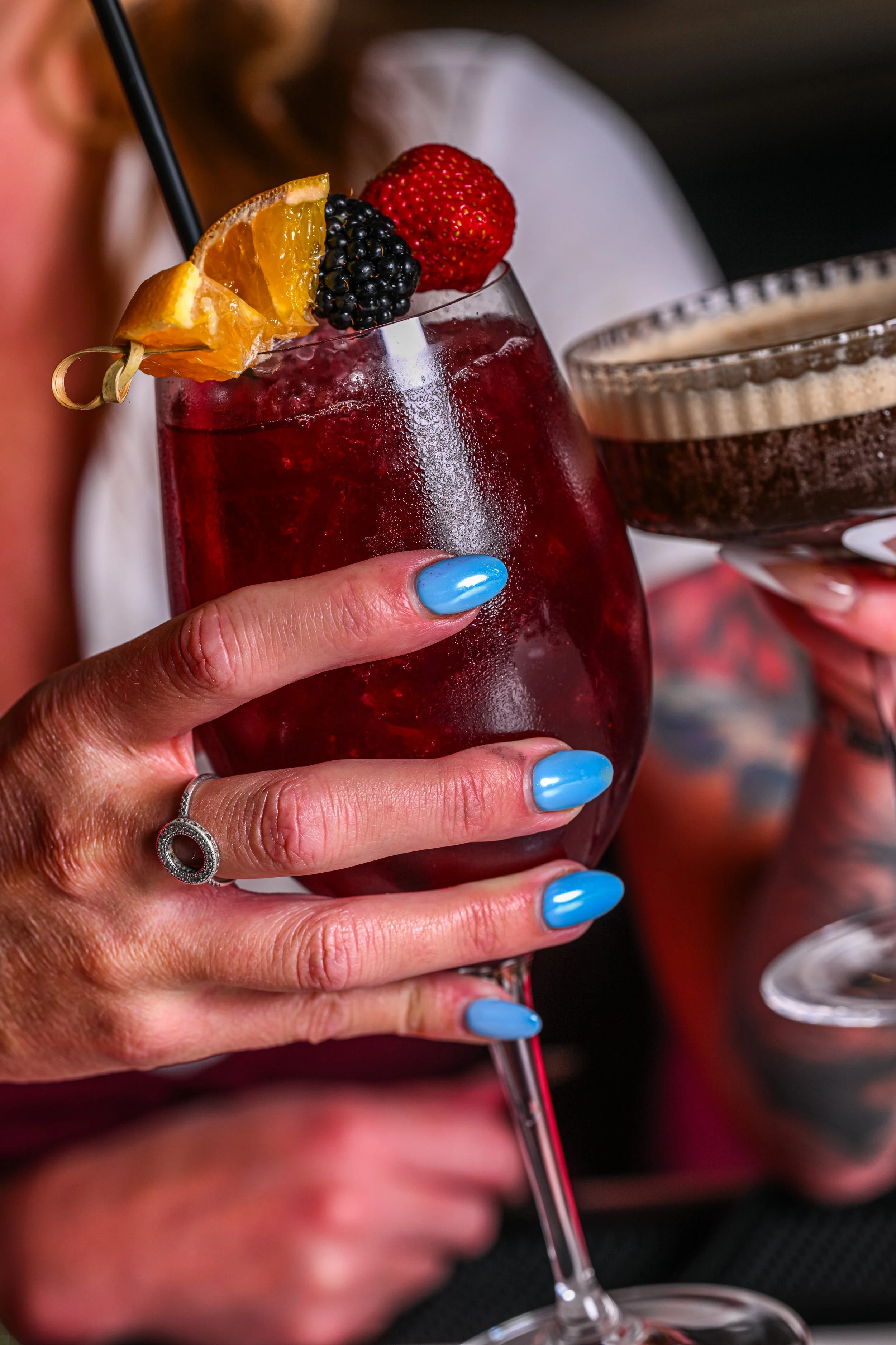 Person holding a glass of red cocktail garnished with a strawberry, blackberry, and orange slice, with another glass of a layered drink visible in the background.