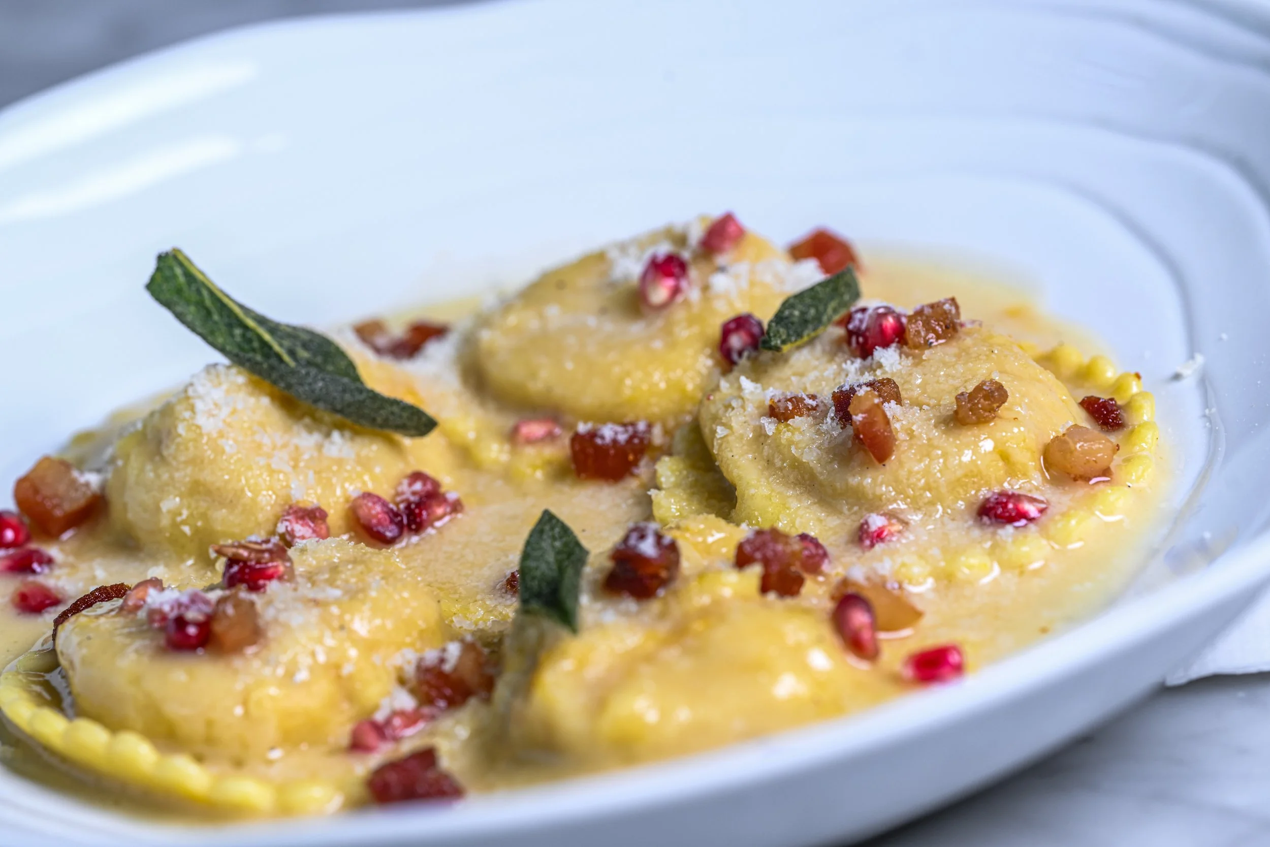 Close-up of gnocchi in a creamy yellow sauce, garnished with red pomegranate seeds and small leaves, served on a white dish.