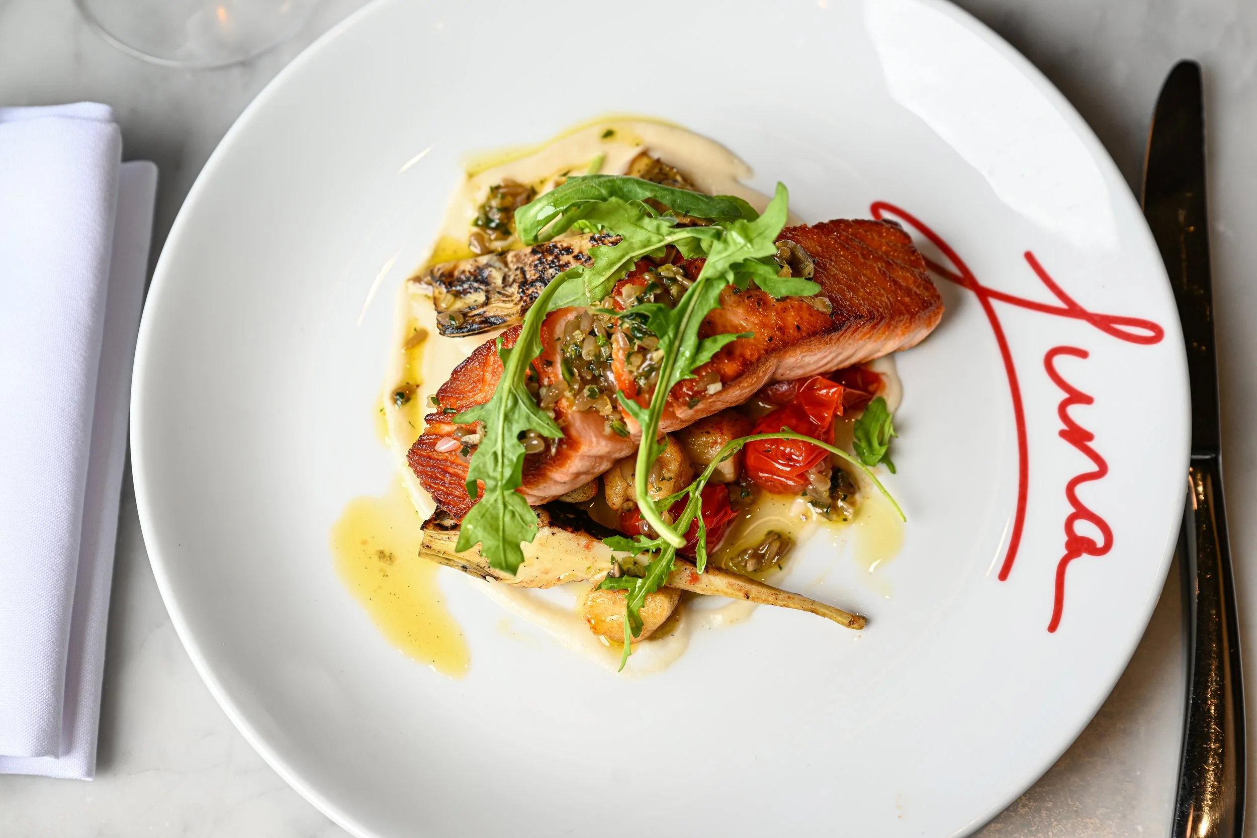 A plated gourmet dish featuring a cooked salmon fillet topped with herbs and vegetables, garnished with fresh greens, served on a white plate with red script writing, accompanied by grilled vegetables and drizzled with sauce.