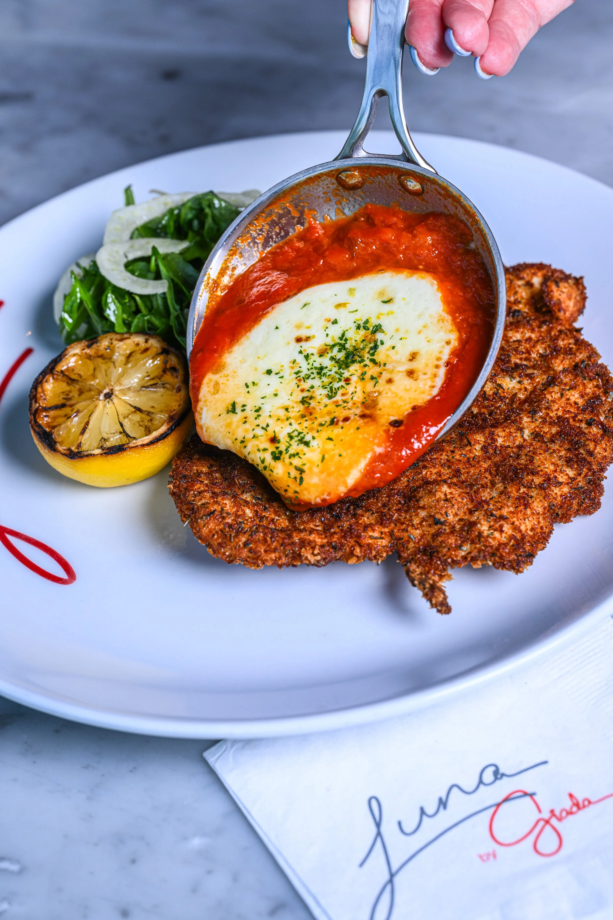Fried chicken cutlet topped with tomato sauce and melted cheese, served with a side of sautéed greens and grilled lemon, on a white plate with a paper napkin nearby.