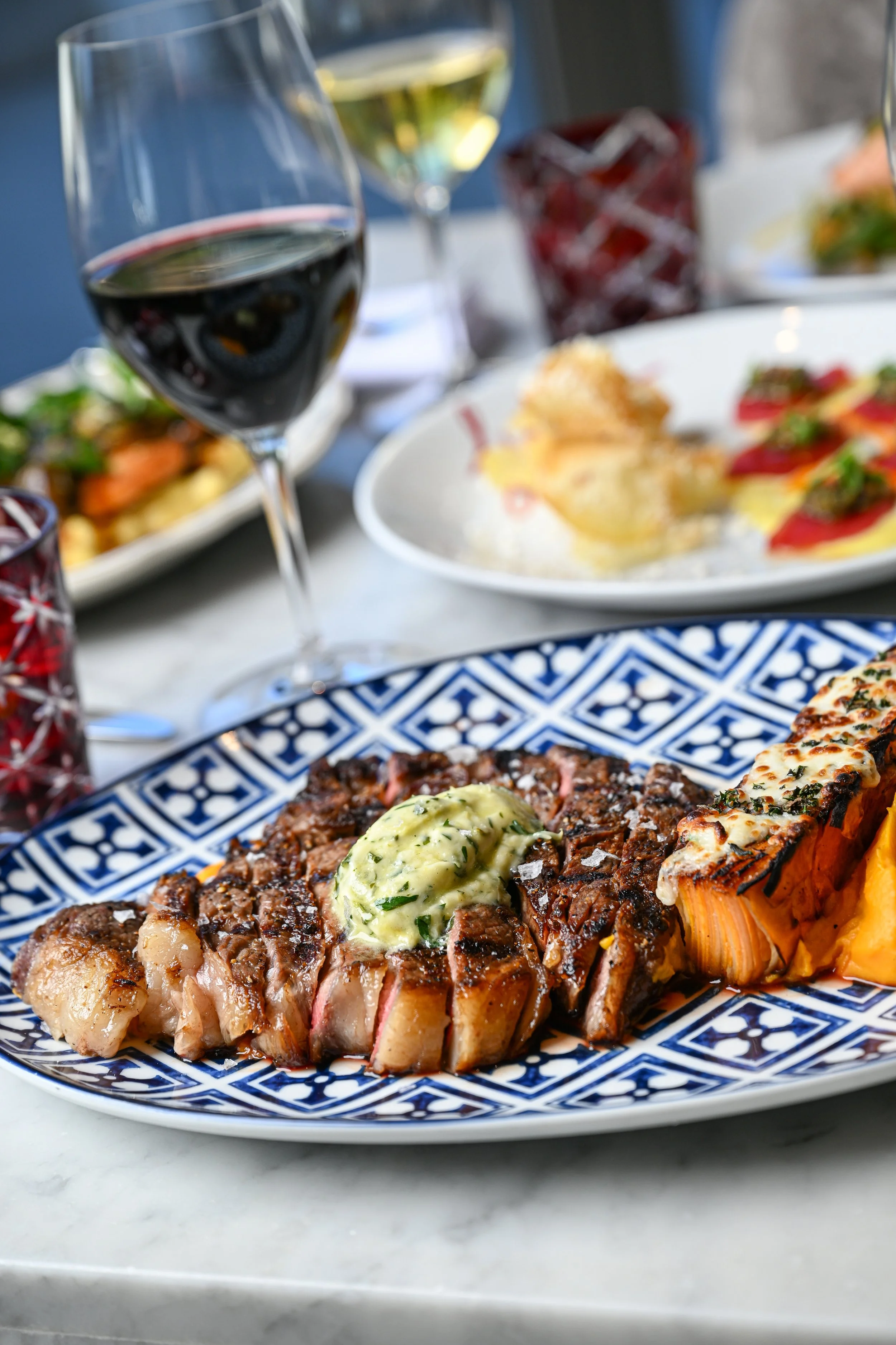 A plate with a cooked steak topped with herb butter and a crusted lobster tail, with side dishes, glasses of white and red wine, and other plates of food in the background on a dining table.