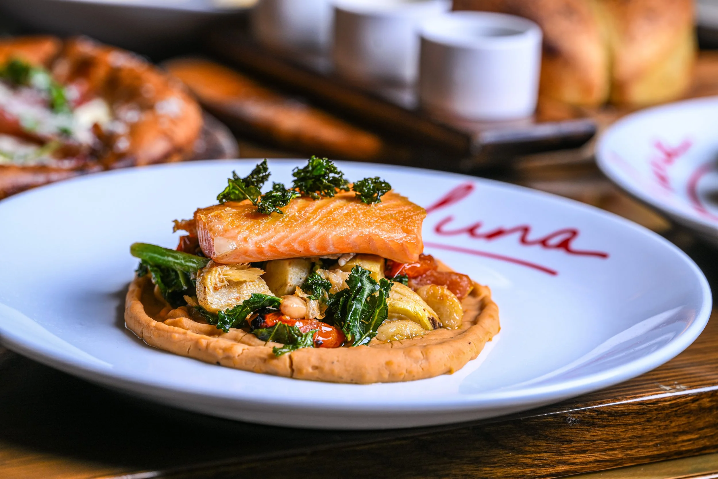 A salmon and vegetable tartlet on a white plate with a red logo, garnished with herbs, with a blurred background of additional dishes and cups on a wooden surface.