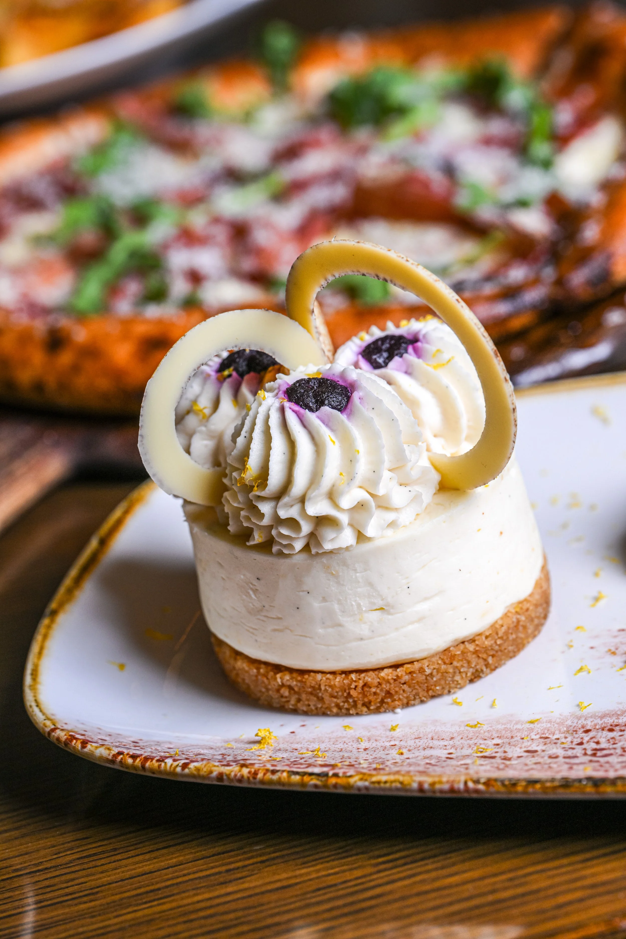 A small dessert with a round biscuit base topped with whipped cream, garnished with blackberries, white and dark chocolate curl decorations, served on a decorated ceramic plate.