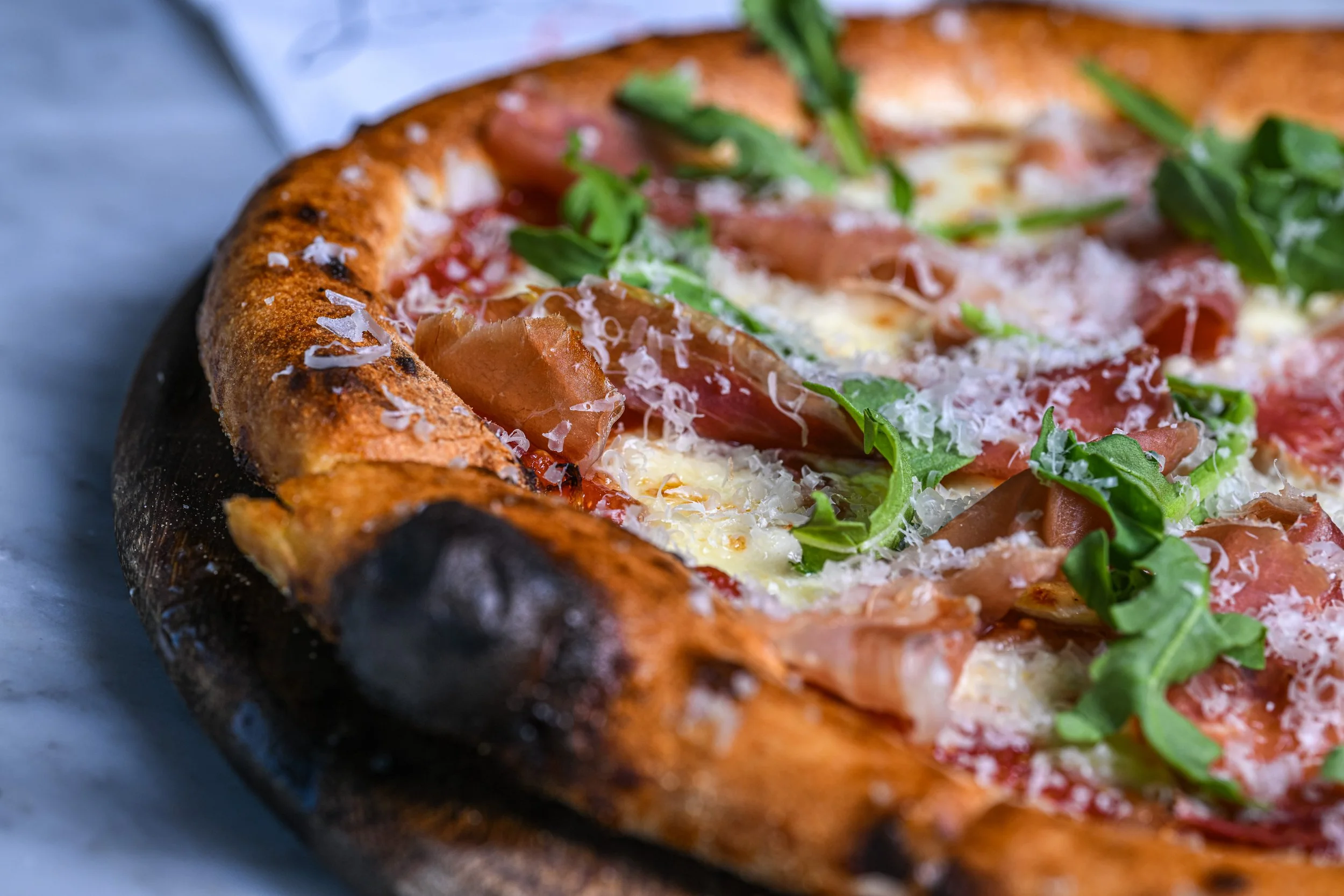 Close-up of a pizza with crispy crust, topped with slices of tomato, arugula, grated cheese, and prosciutto.