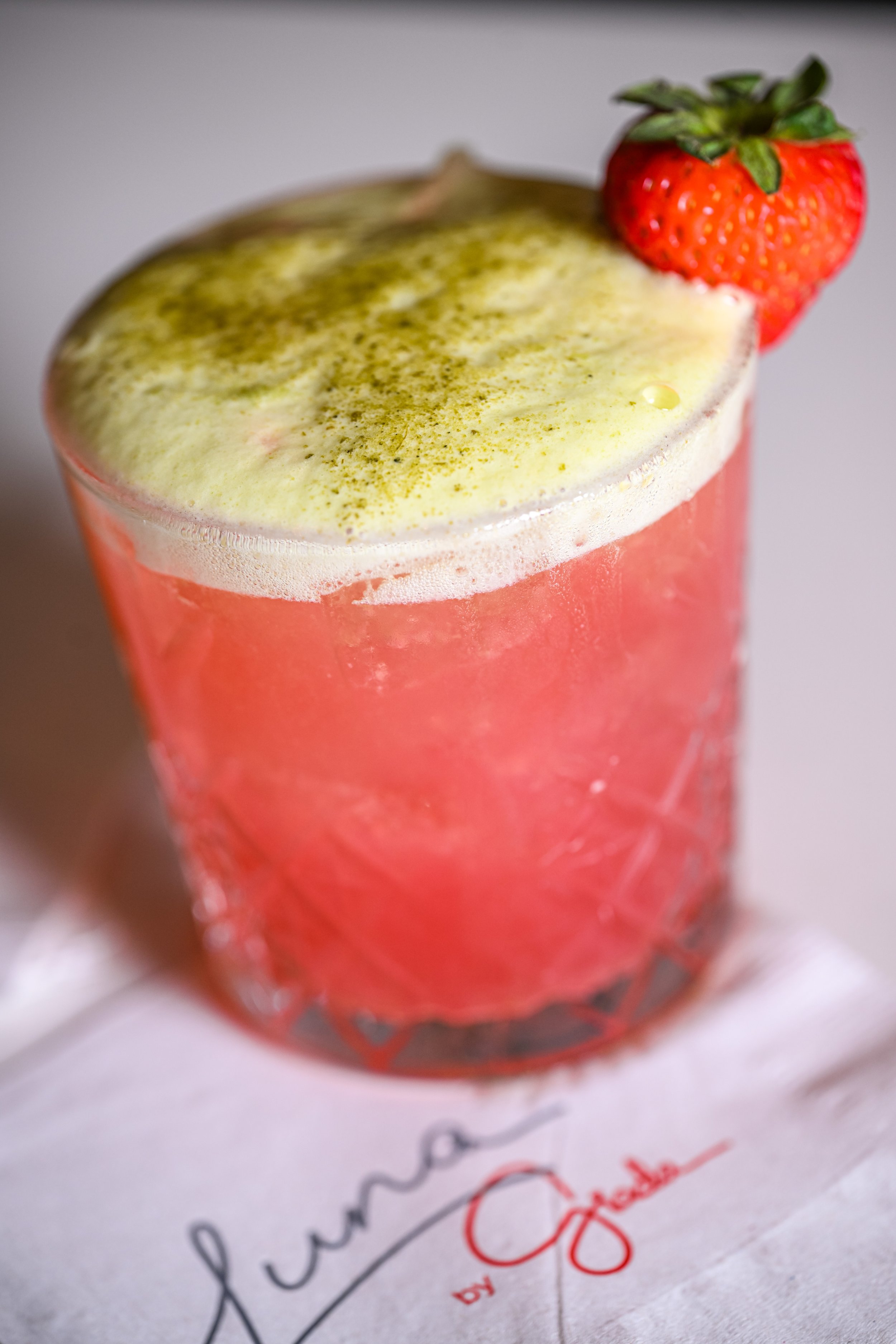 Close-up of a pink strawberry-flavored beverage in a textured glass, topped with a fresh strawberry and dusted with matcha powder.