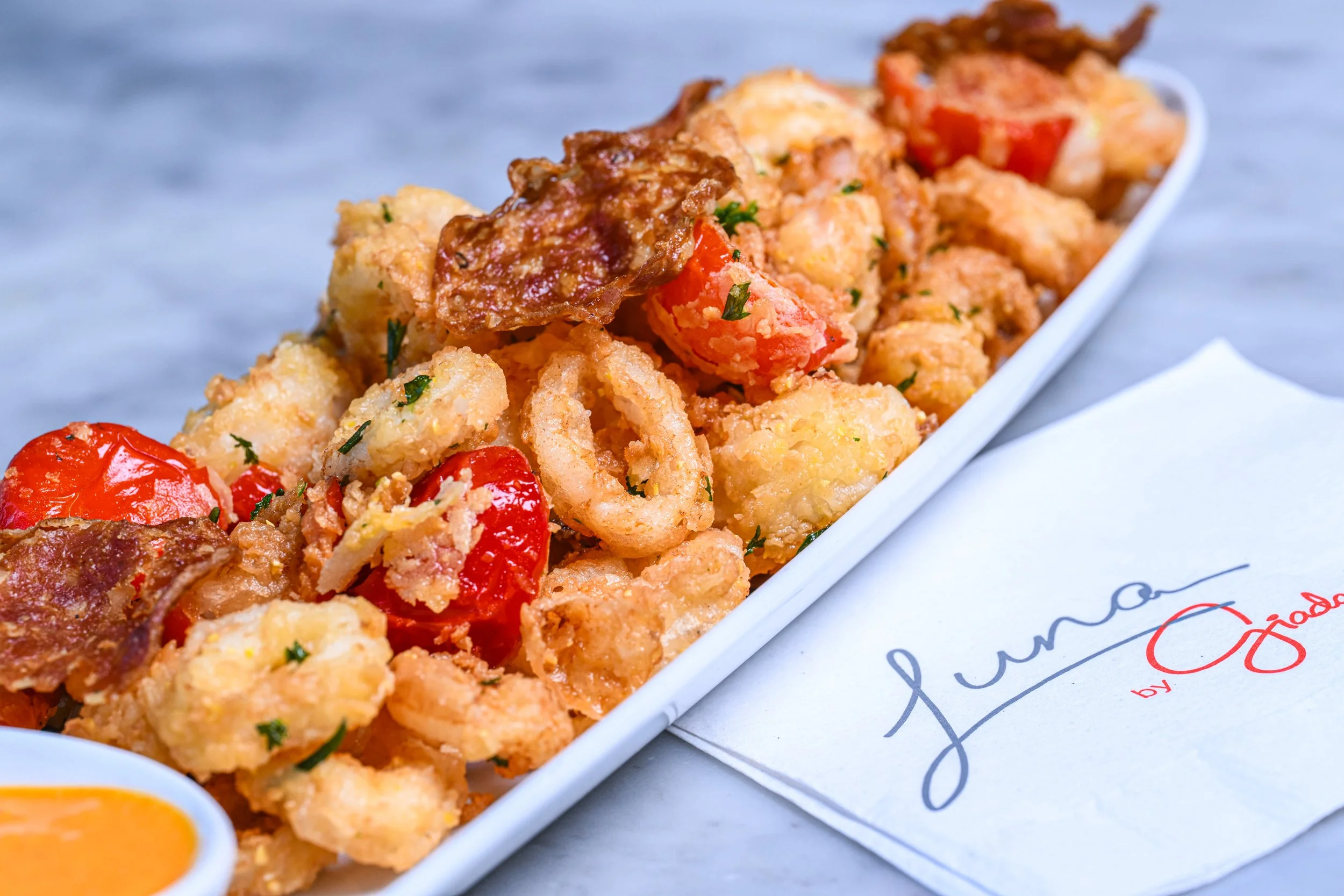 Close-up of fried shrimp with cherry tomatoes, garnished with herbs on a white serving platter, with a dipping sauce in a small bowl nearby.