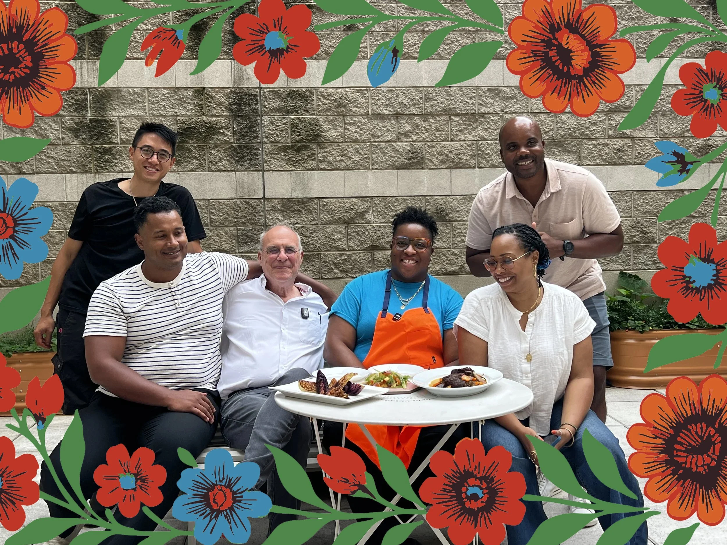 This is a photo of the Community Kitchen team. Six people (two standing and four sitting) behind a table with plates of food. A border of red, blue, and orange flowers (with green leaves and stems) appears at the top and bottom of the photo.