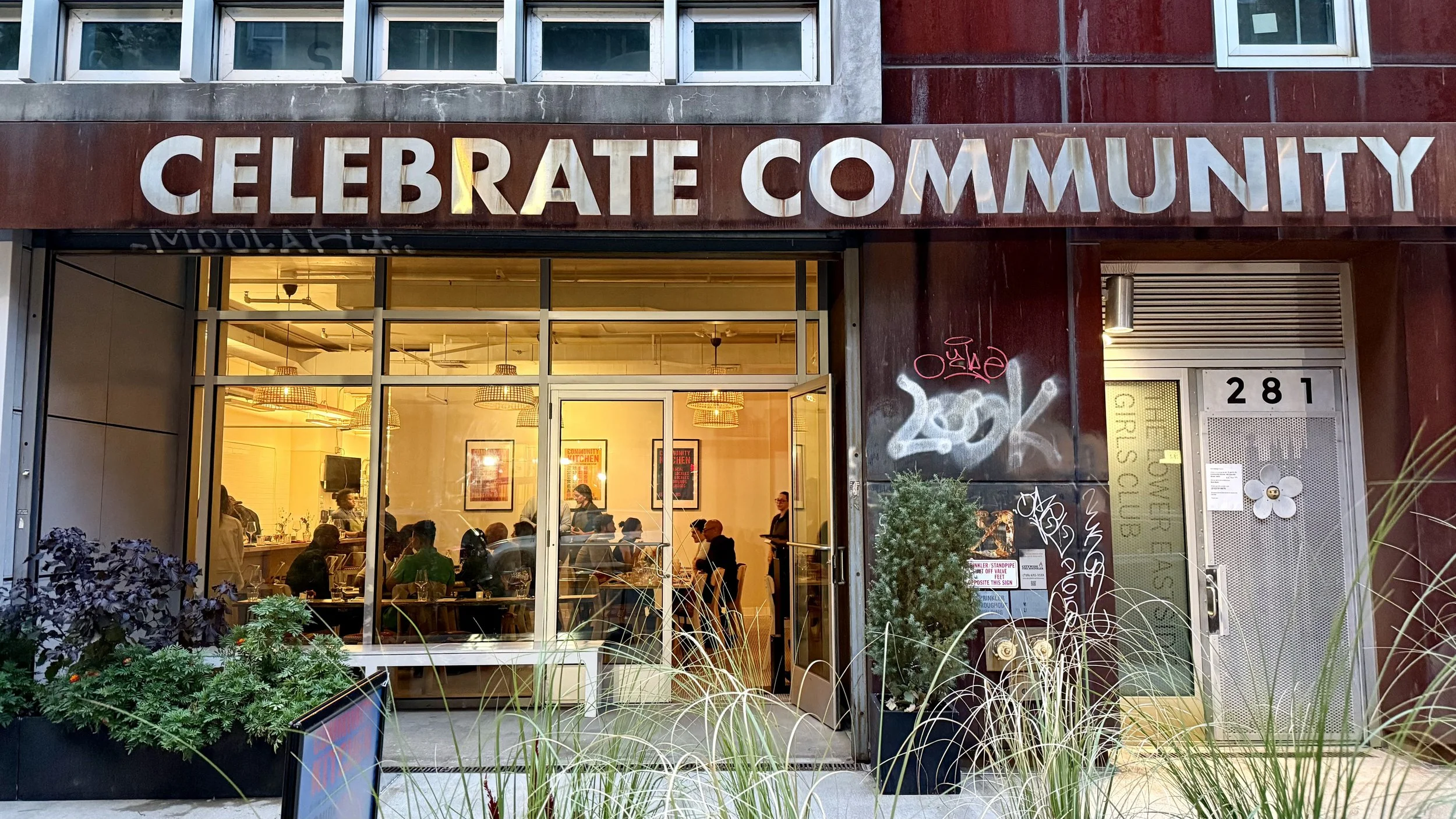 This photo is of the facade of the Community Kitchen pilot restaurant location, which took place at the Lower EastSide Girls Club in NYC. The photo shows a brightly lit restaurant, filled with people, and the facade says "Celebrate Community."