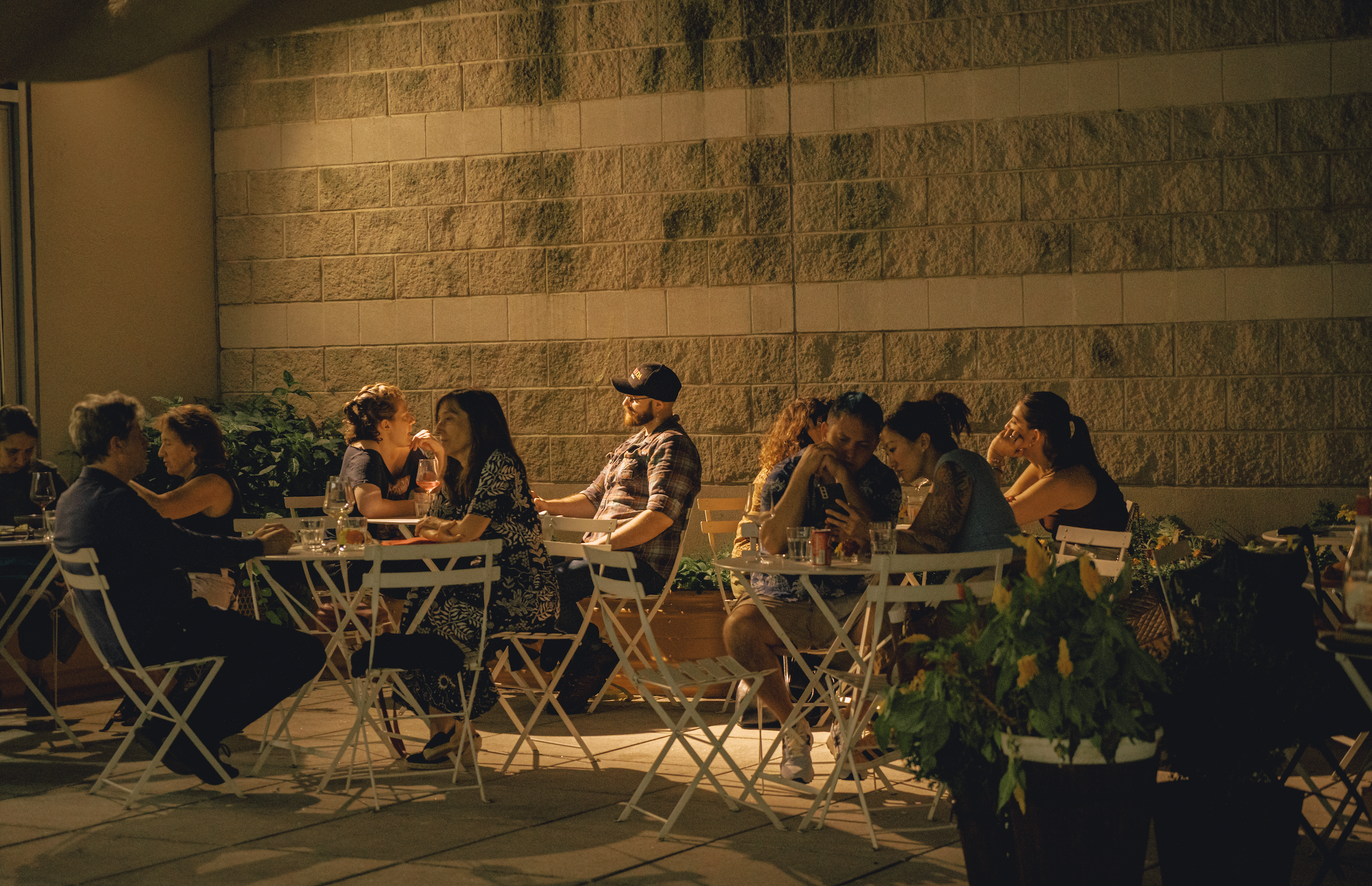 People dining outdoors at night, sitting at white tables with drinks, against a brick wall, illuminated by warm lighting.