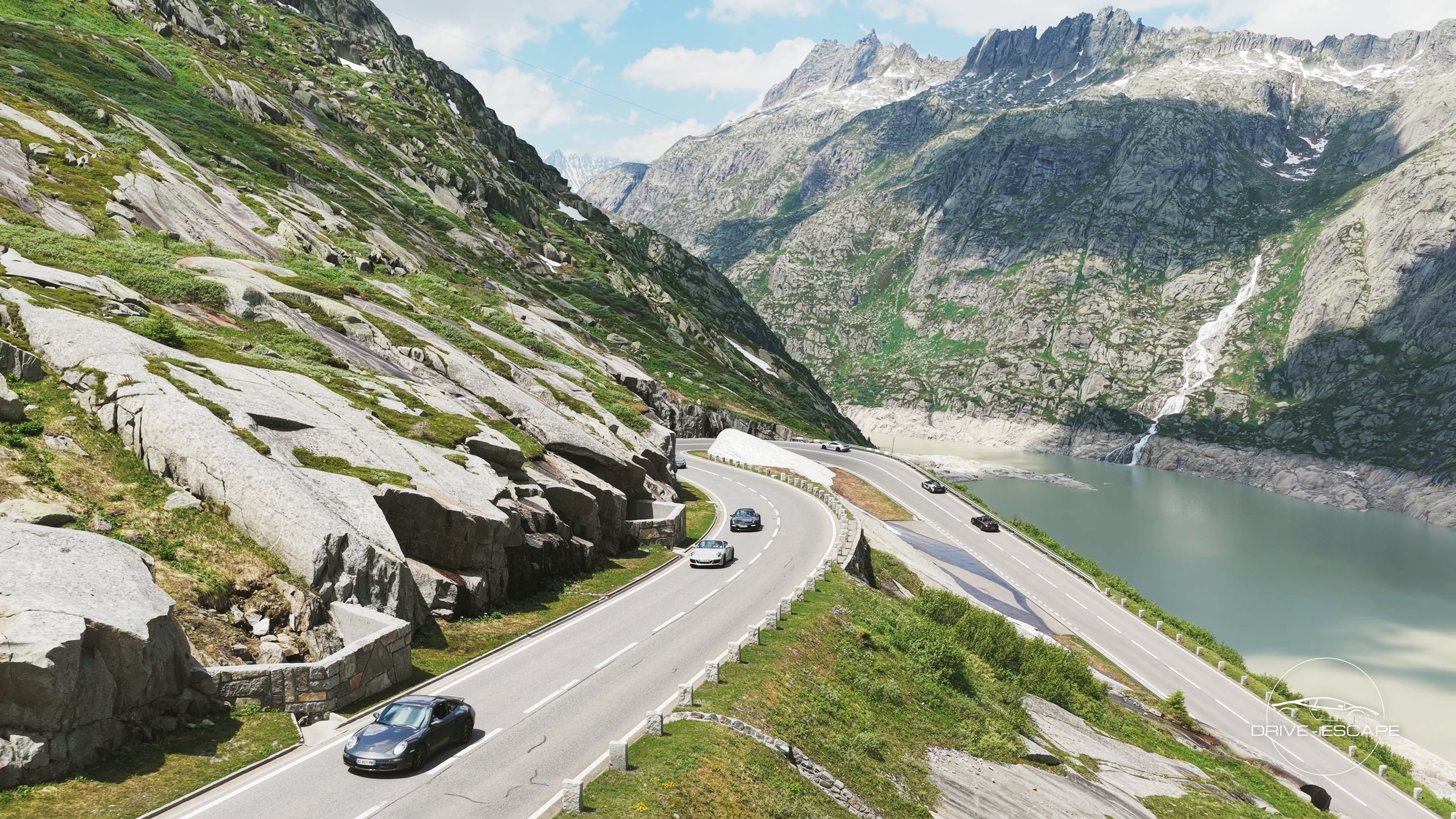 Route de montagne serpentant le long d'un lac, entourée de falaises rocheuses et de montagnes en arrière-plan, avec plusieurs voitures en déplacement.
