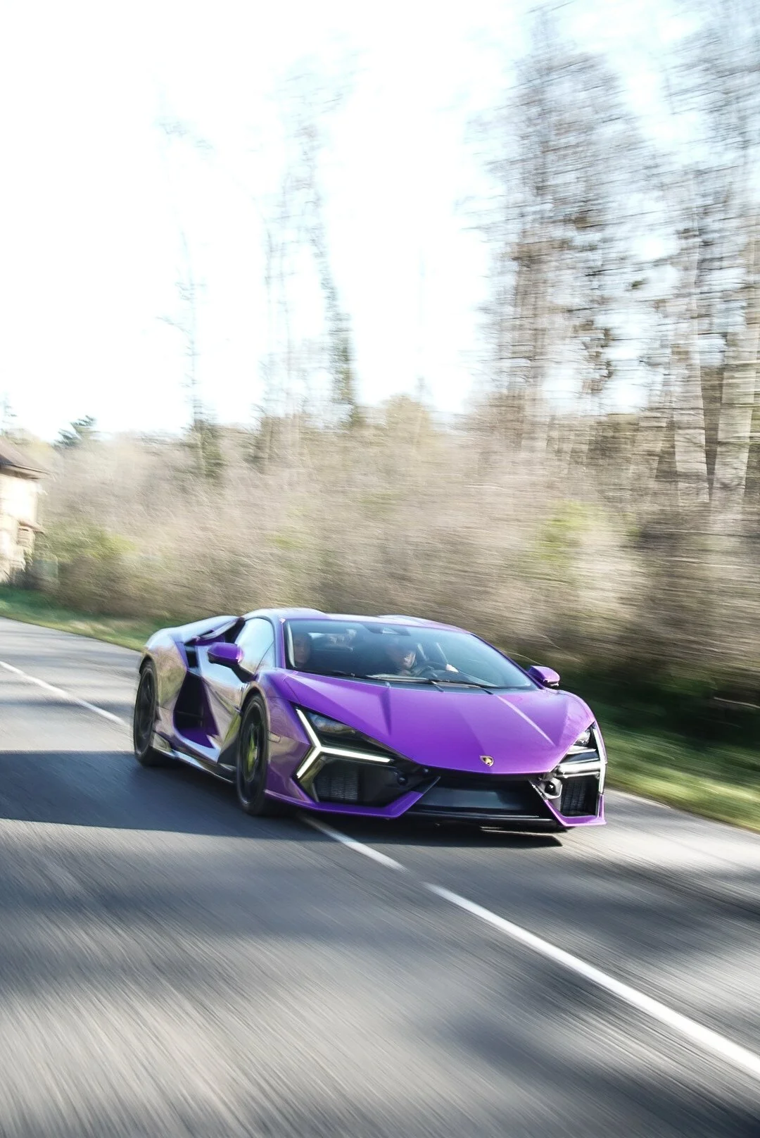 Une voiture de sport violette conduite à grande vitesse sur une route, avec un fond flou d'arbres et de verdure.
