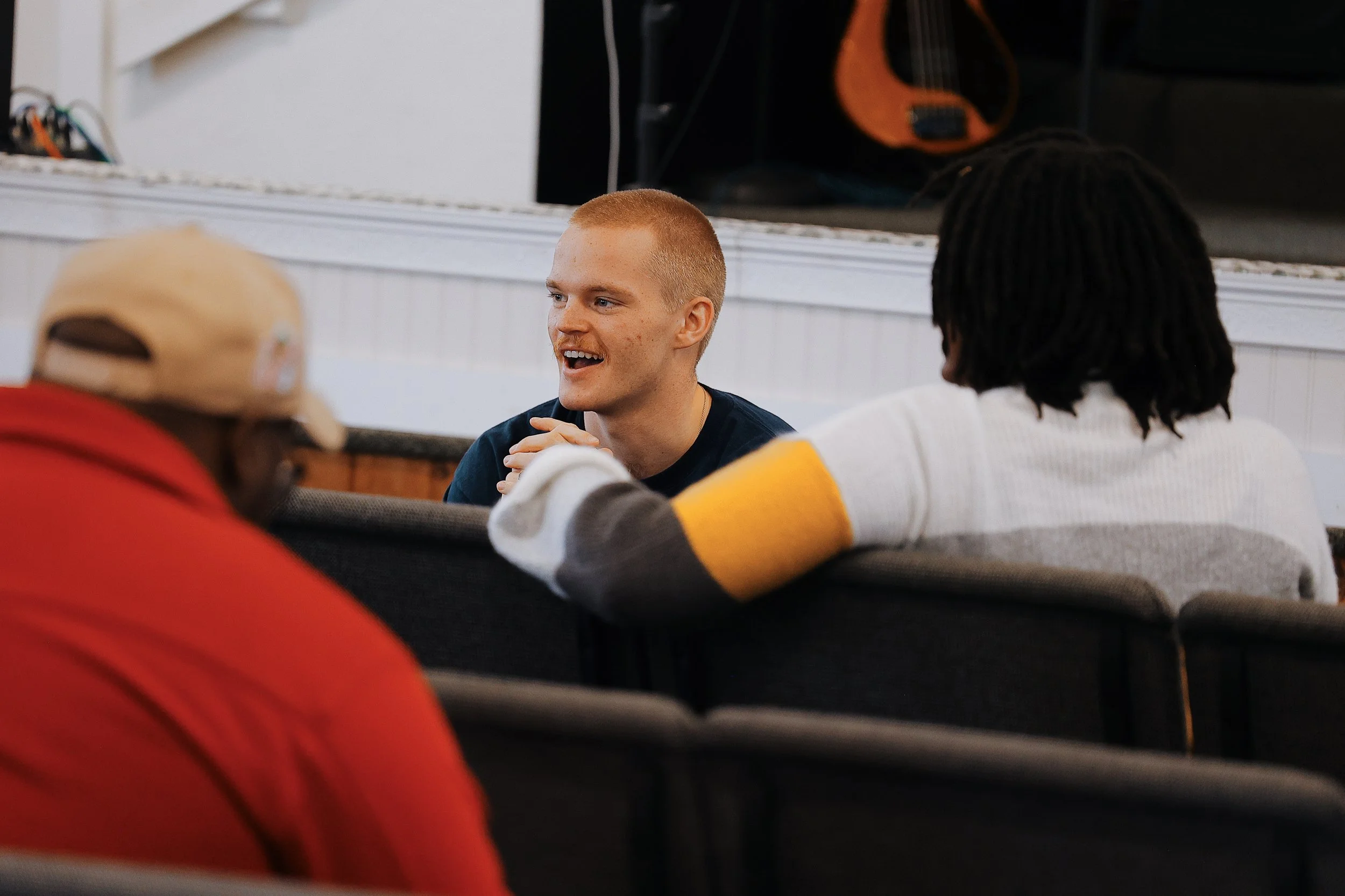 A young man with short blonde hair and a big smile is talking to two other individuals in a casual indoor setting. One person wears a beige cap and a red jacket, and the other has black curly hair and wears a white shirt with yellow accents. A guitar is visible in the background.