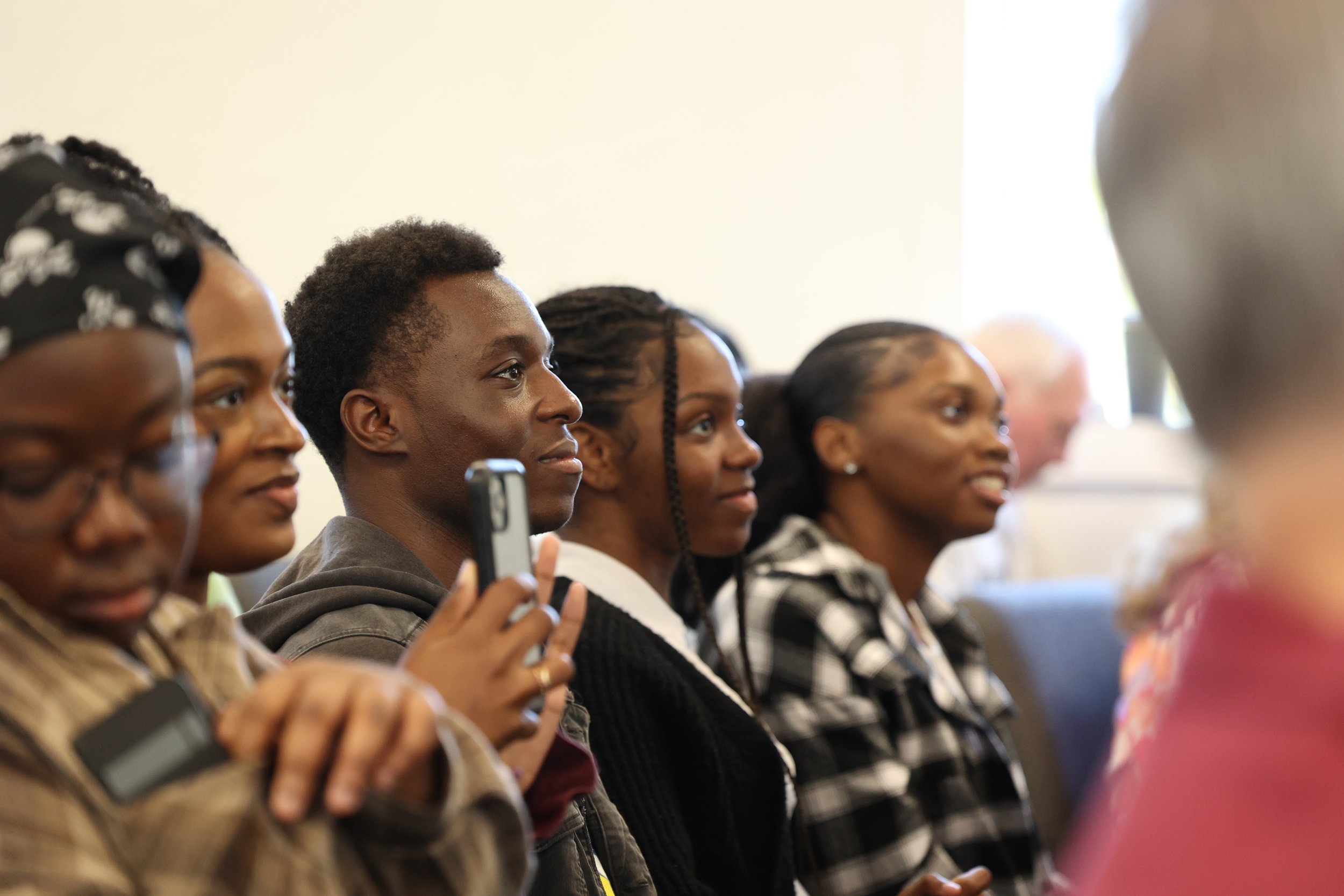 A group of diverse people sitting in a row, attentively listening in a seminar or classroom setting, some taking photos or notes.