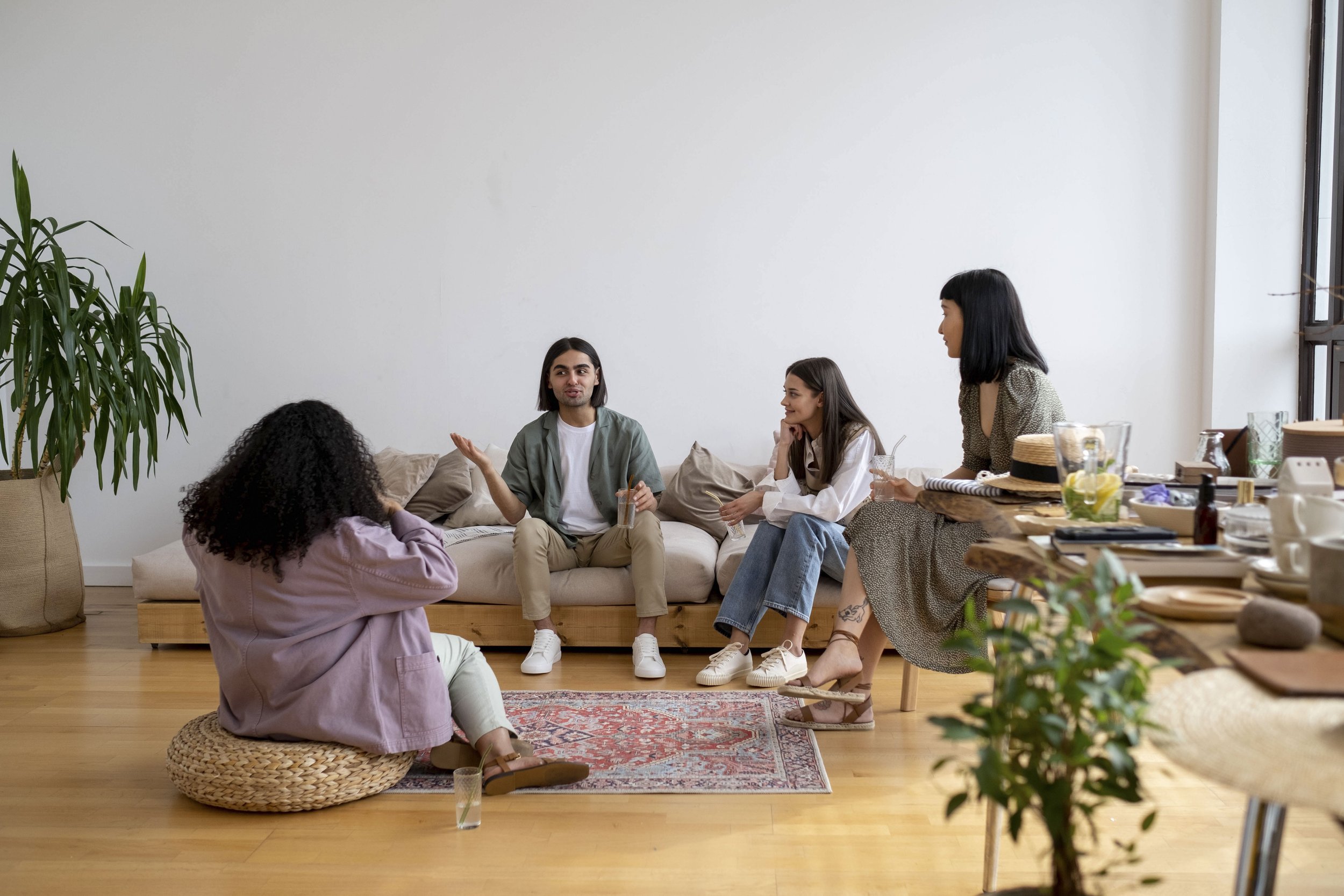 A diverse group of adults sit on a couch, chairs, and floor cushions, while in conversation with each other. A table with food and notebooks is visible in the corner of the room, framed by plants.