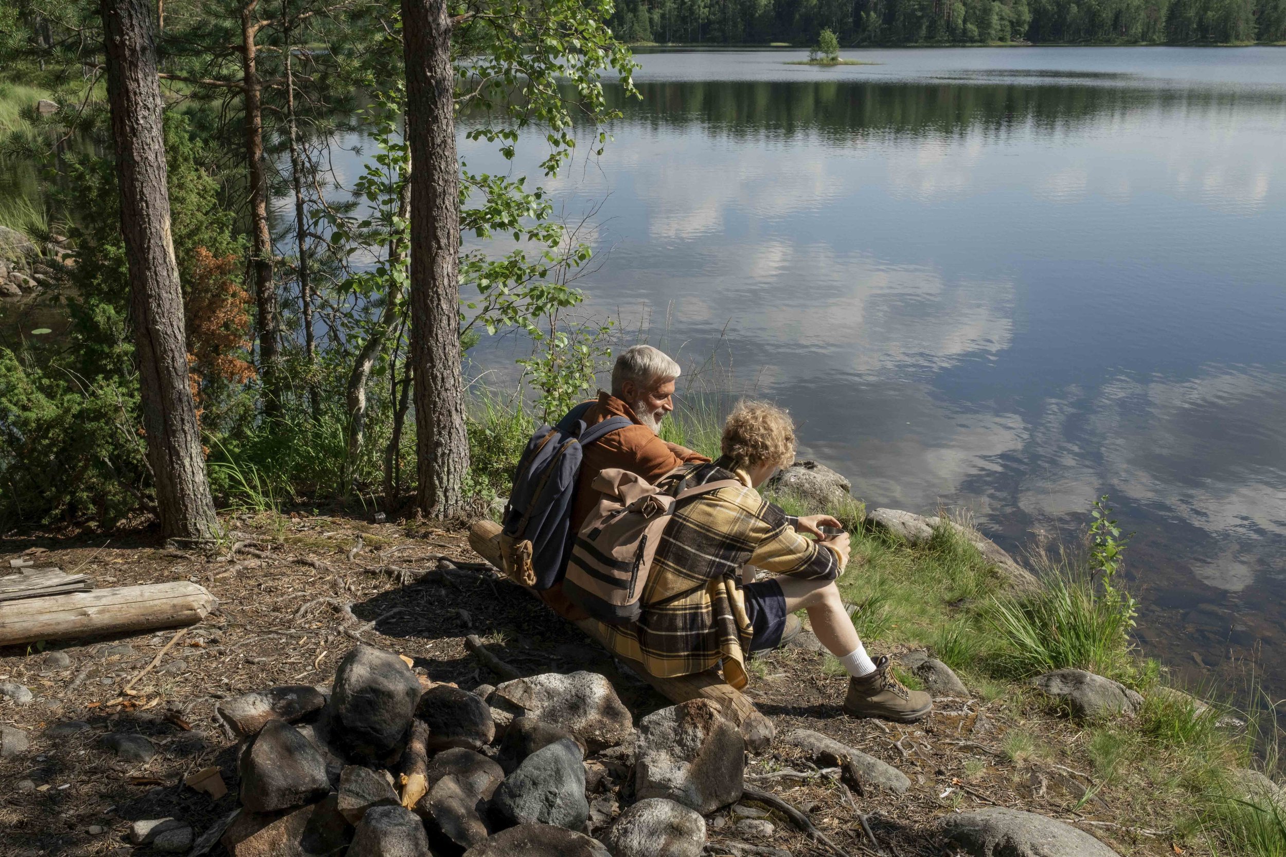A father and son sit outside, near the edge of a body of water in front of them, trees surrounding them. A fire pit made of found rocks is visible behind them, and they are wearing backpacks and hiking boots.