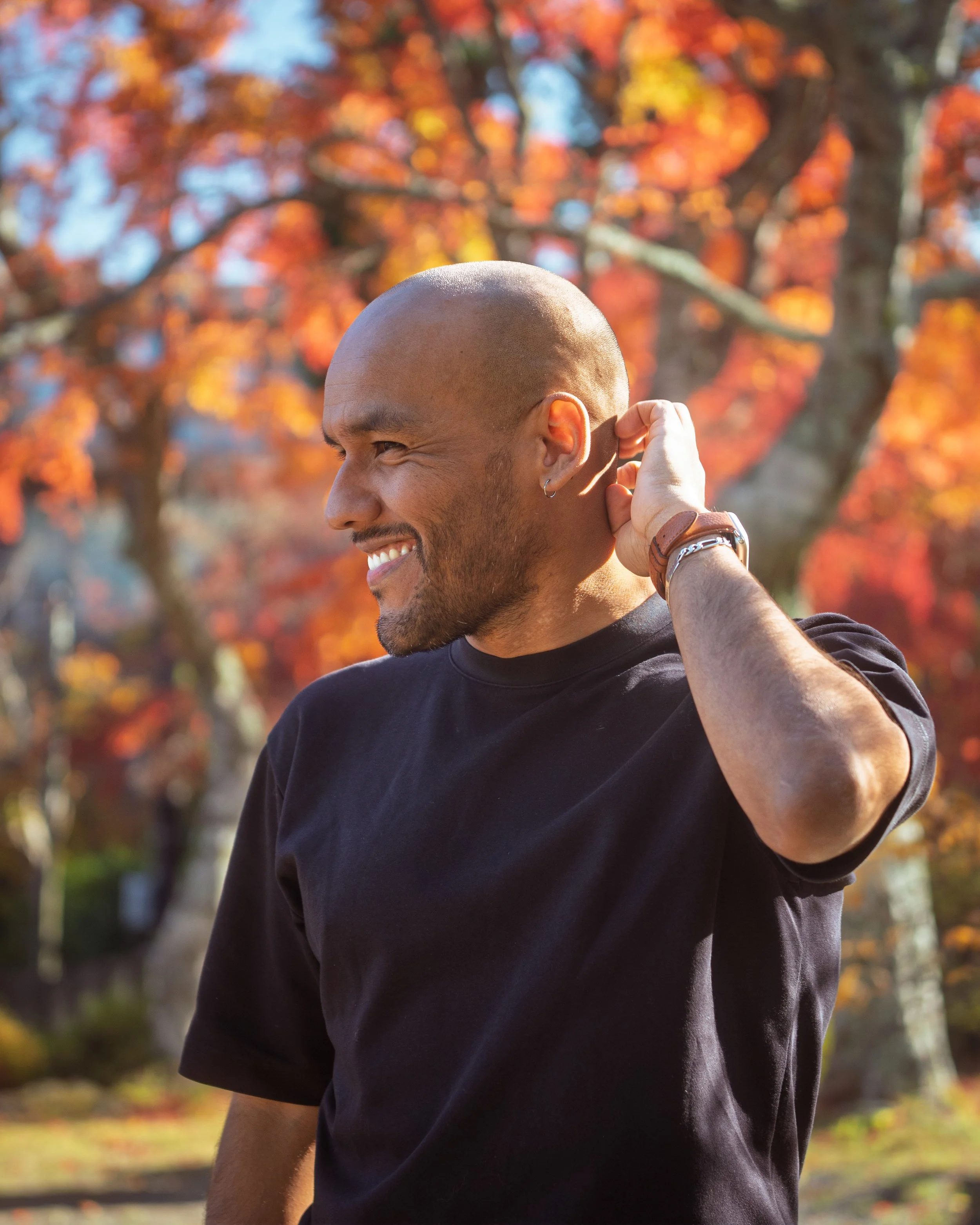Smiling man with a shaved head and beard outdoors, touching his ear, with colorful fall foliage in the background.